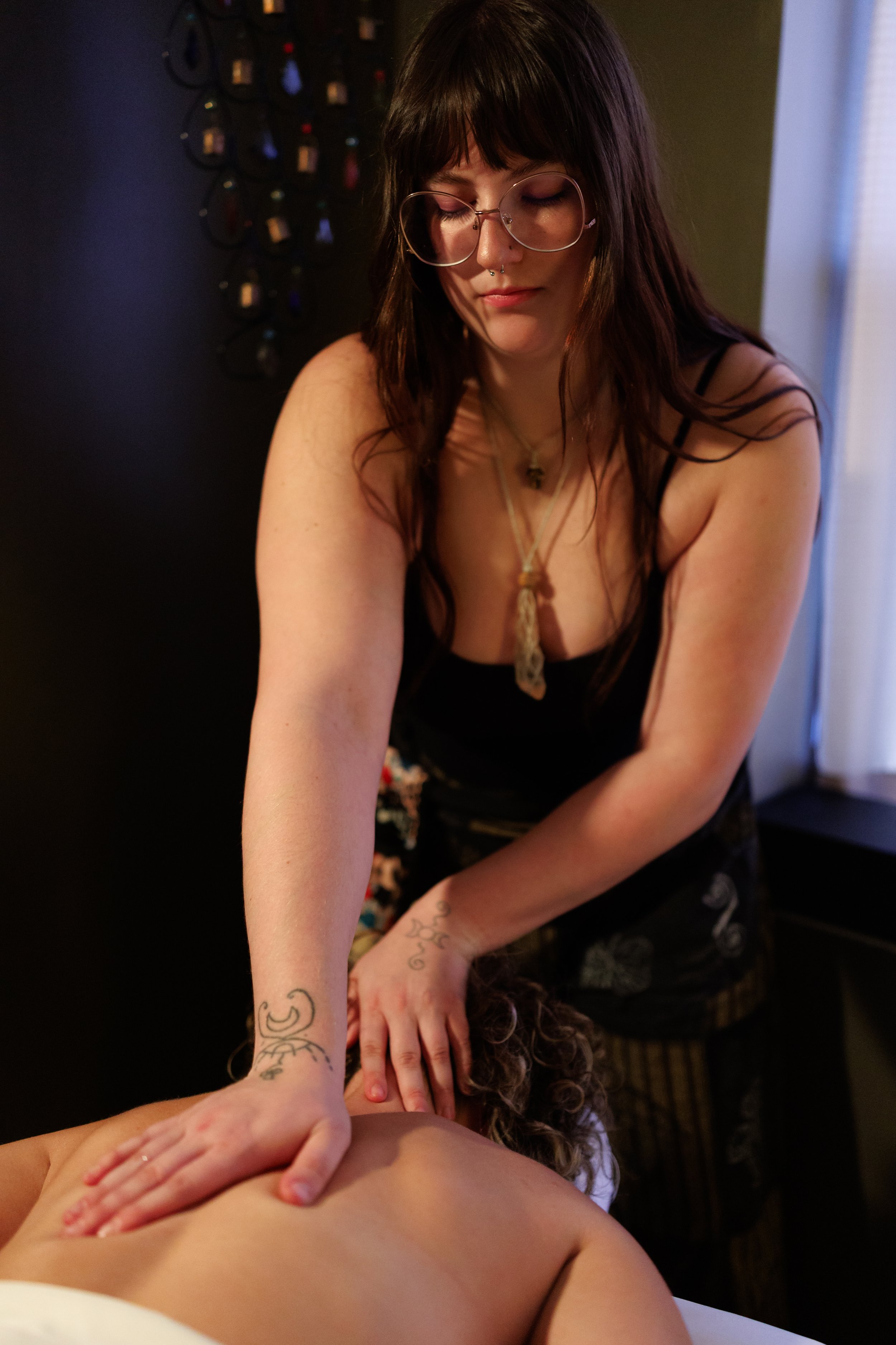 A intelligent woman with dark hair and glasses giving a massage to a person lying face down, with her hands on their back. The setting appears to be indoors with warm lighting. A cozy and comfortable vibe.