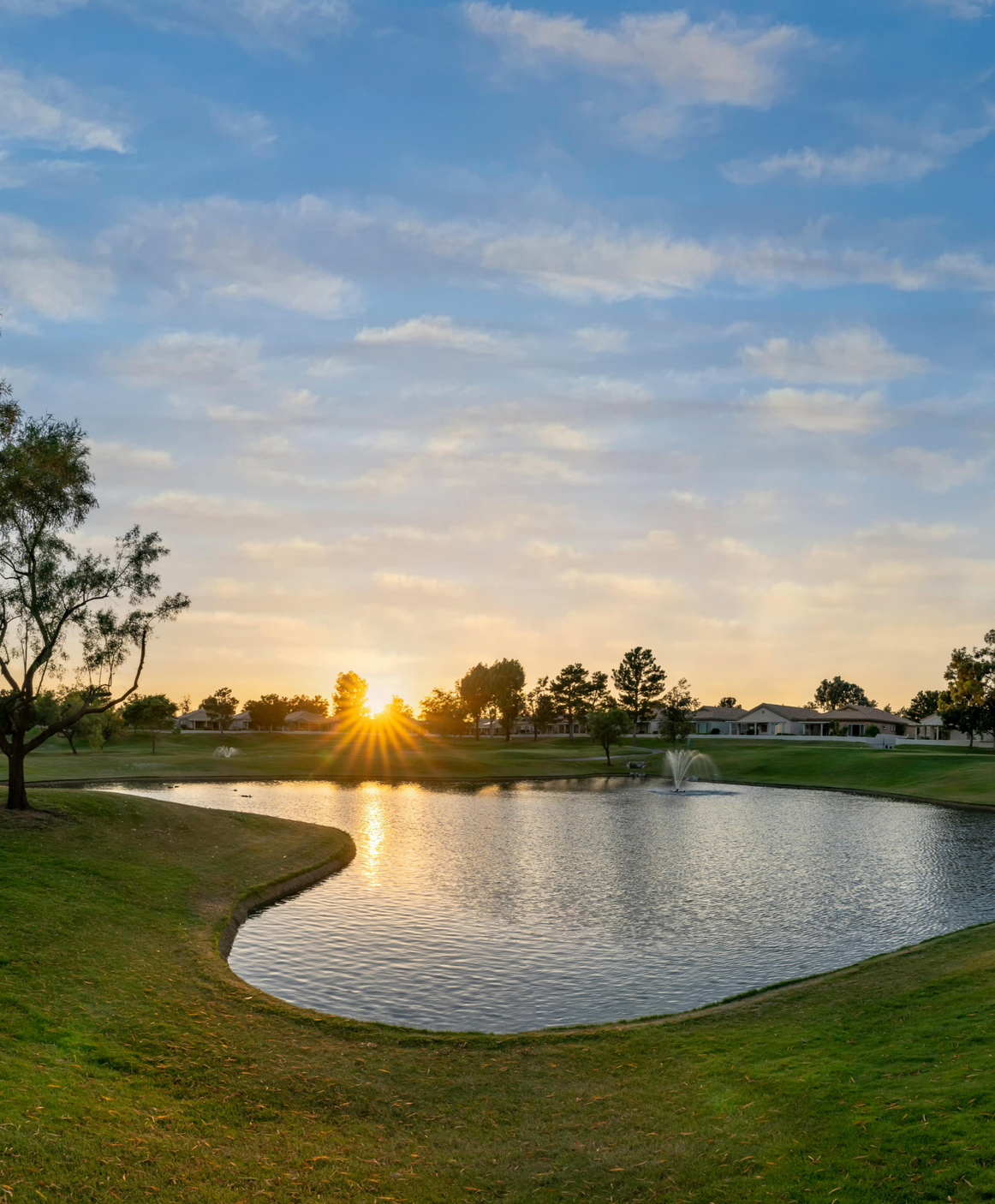 A serene lake at sunset with fountain, surrounded by green grass, trees, and residential houses in the background, under a partly cloudy sky.