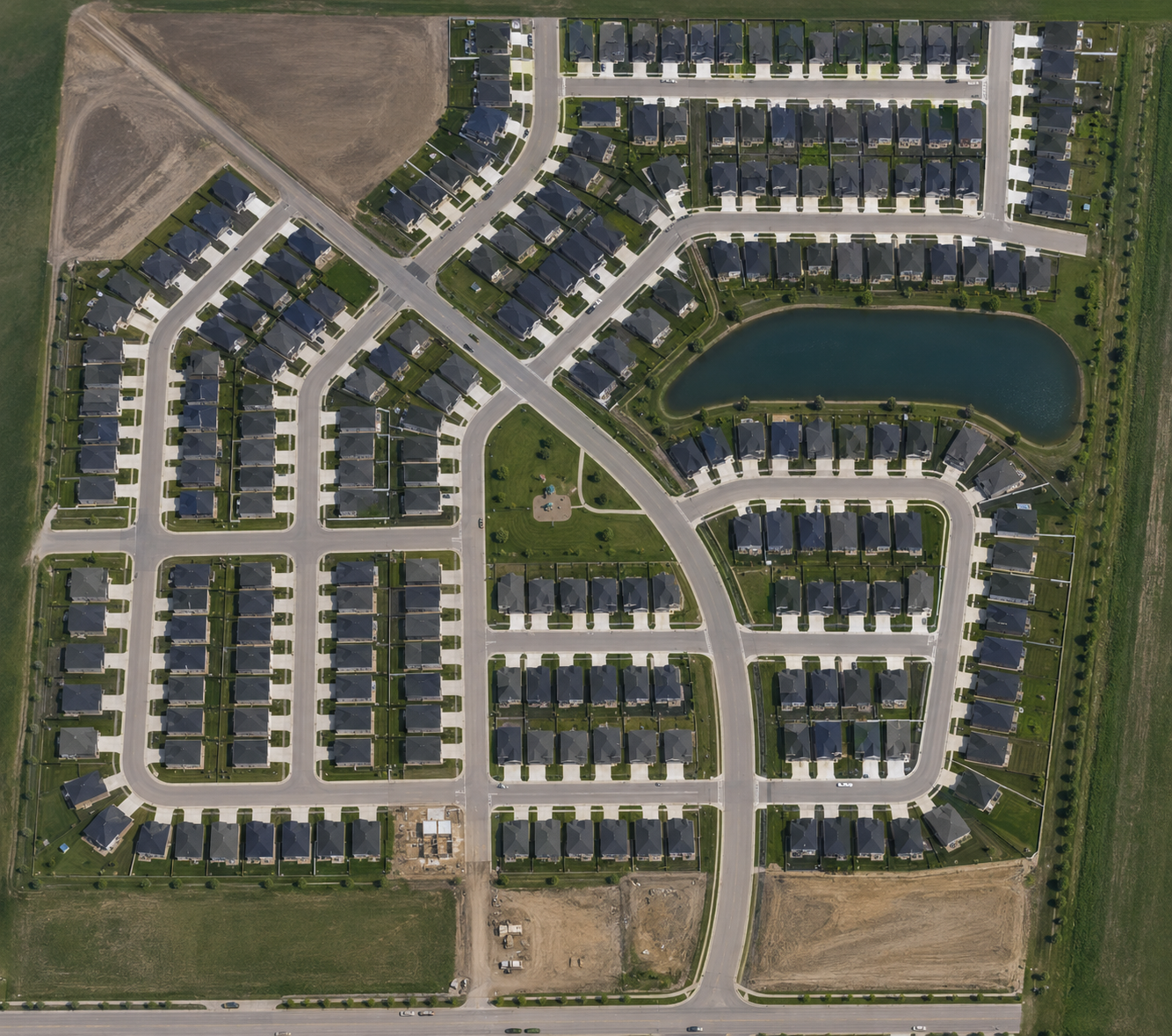 Aerial view of a residential neighborhood with houses, roads, green lawns, a pond, and adjacent open fields.