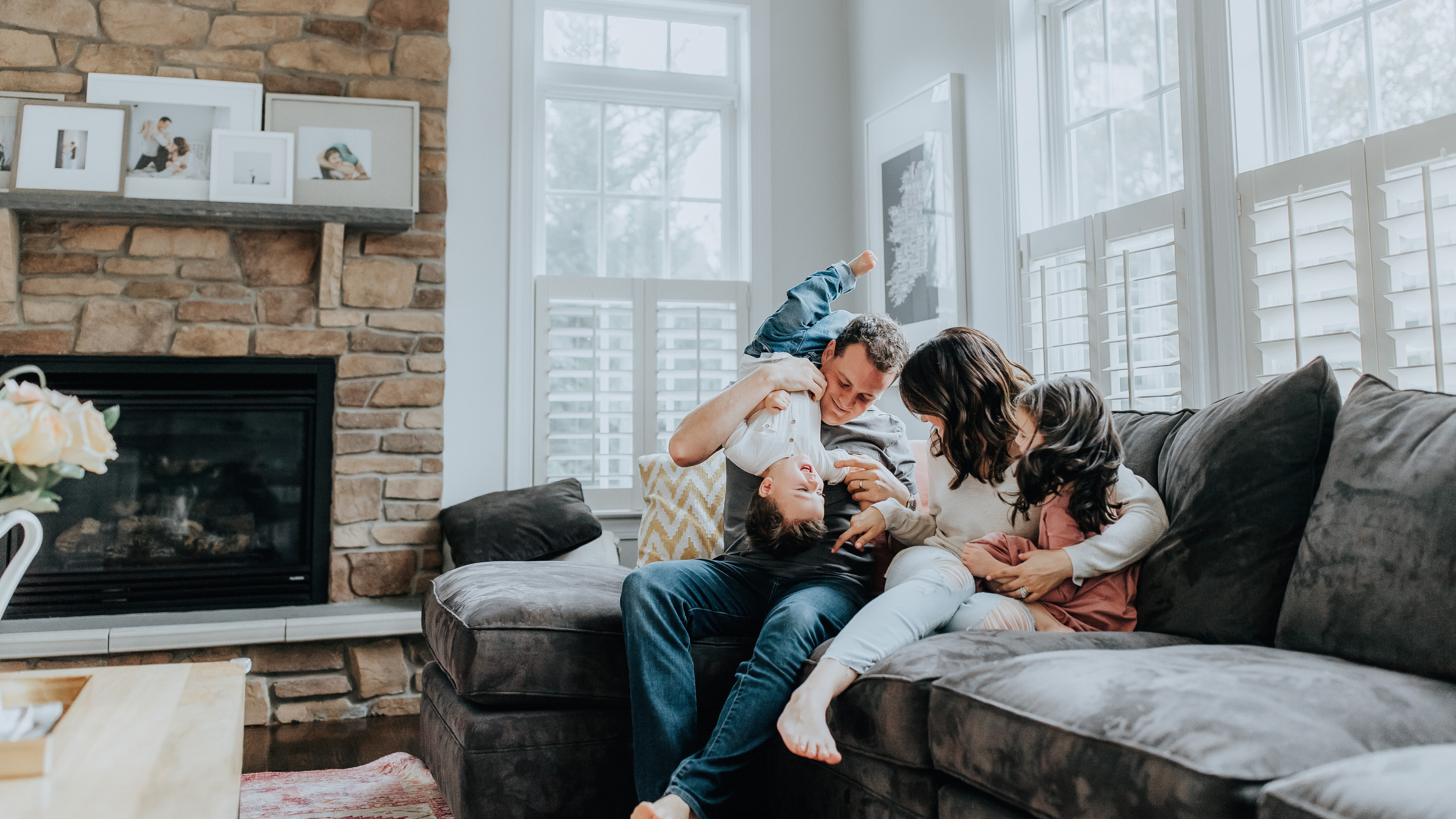 A family of four playing and laughing together on a comfortable sofa in a brightly lit living room with large windows and a brick fireplace.