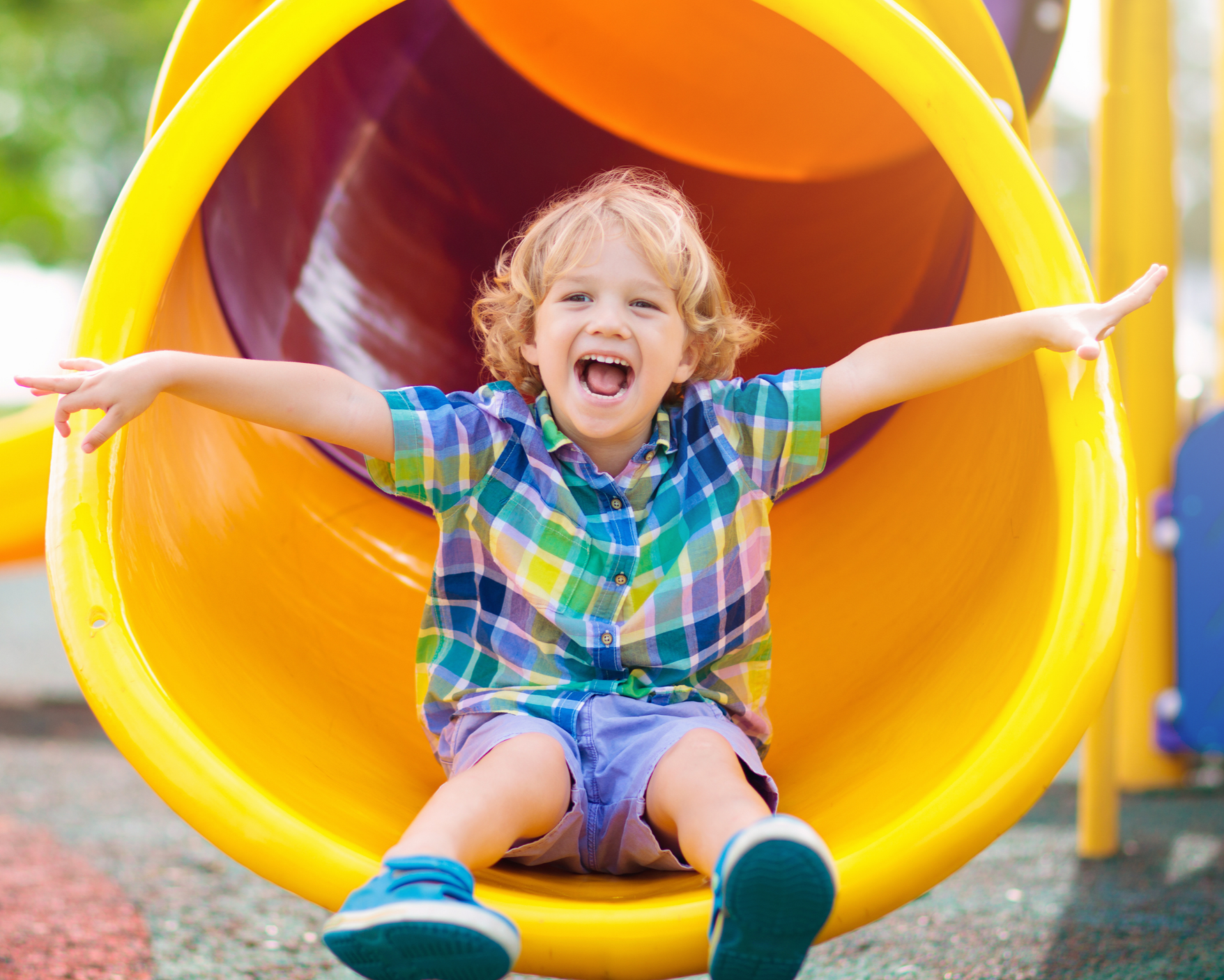 A young boy with blonde hair in a plaid shirt is exiting a yellow playground slide with a big smile and arms outstretched.