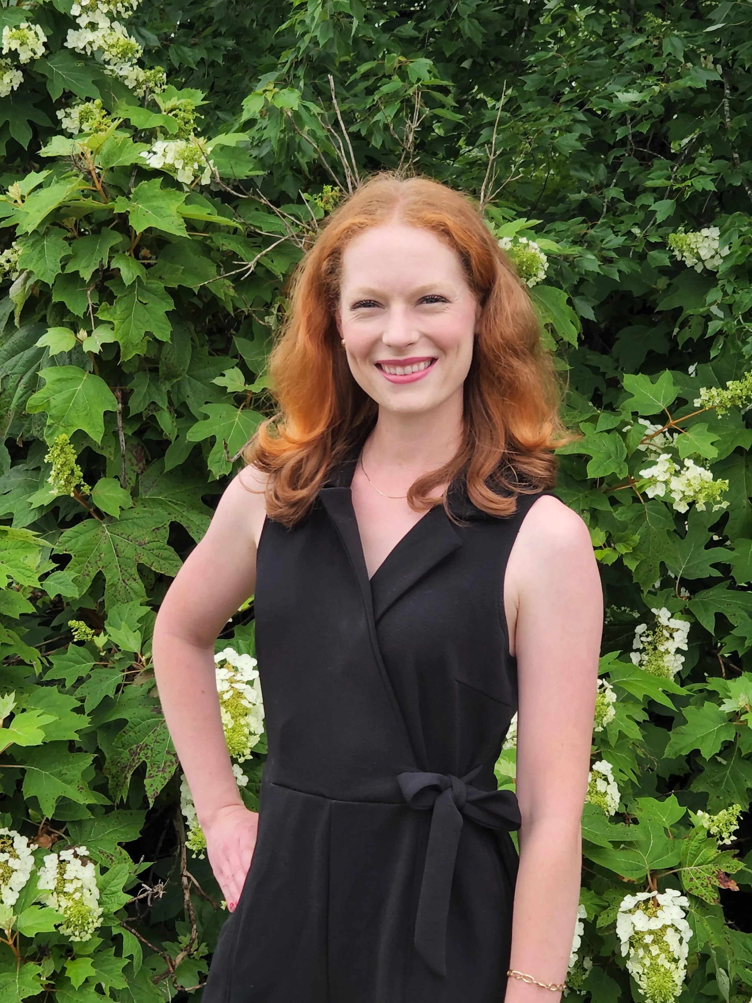 A woman with red hair smiling in front of green leafy plants and white flowers.