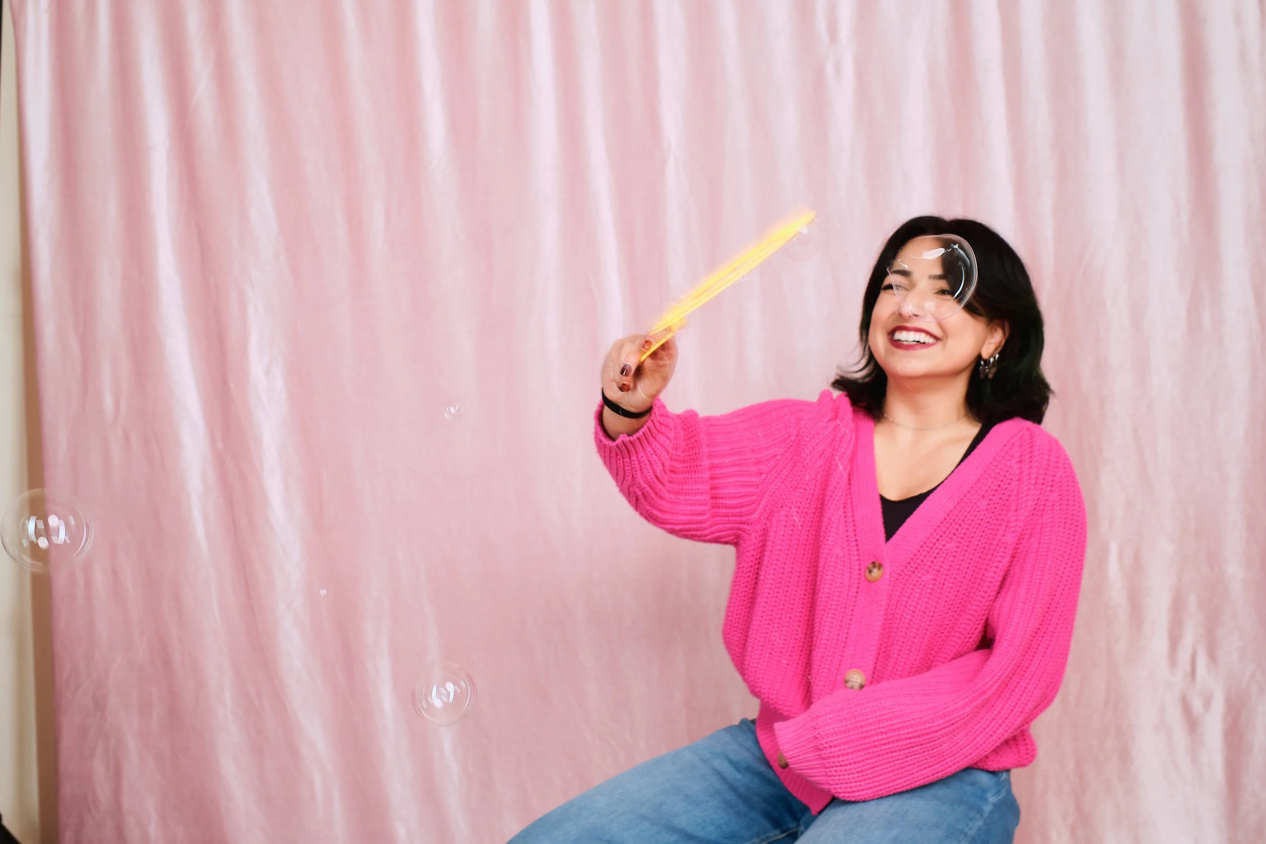 Woman smiling and blowing bubbles in front of a pink curtain, wearing a bright pink sweater and blue jeans.
