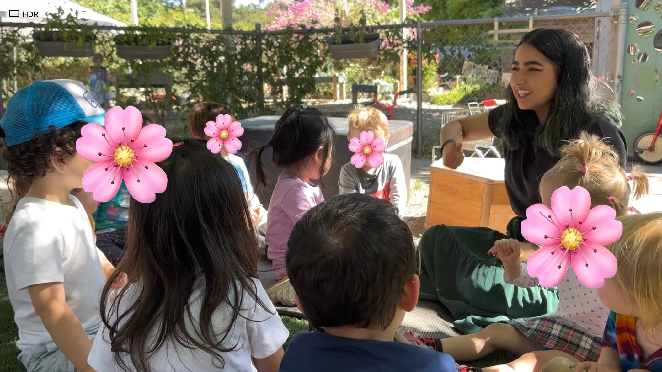 A group of children gathered outdoors, interacting with a woman who is smiling and leaning forward. The children have covers over their faces, with some obscured by pink flower illustrations. The setting appears to be a playground or park with trees, a fence, and outdoor play equipment in the background.