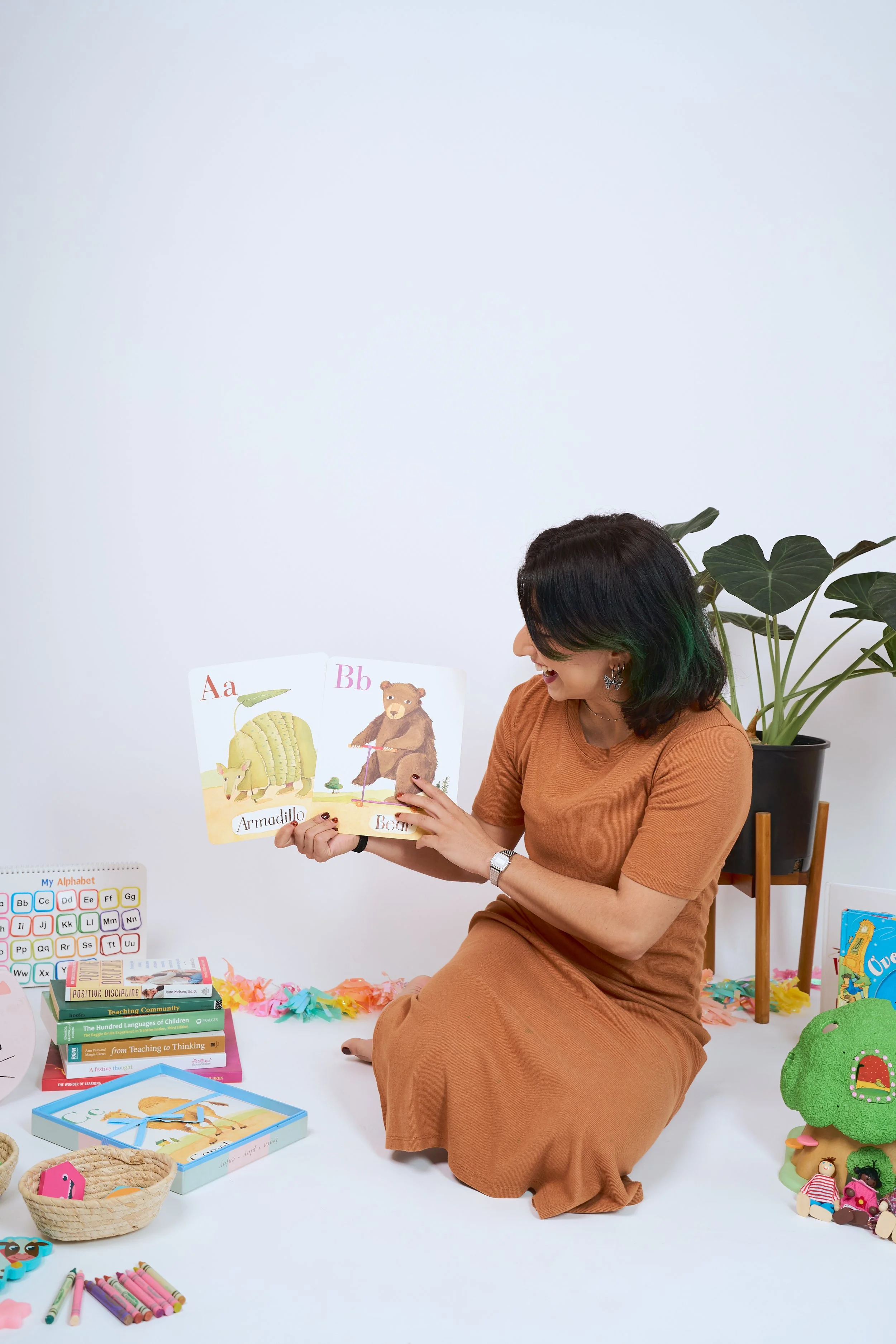 A woman with shoulder-length dark hair and earrings, wearing a rust-colored dress, sitting on the floor and looking at children's books. There are educational materials, books, and toys arranged around her, with a potted plant behind her and a white wall in the background.