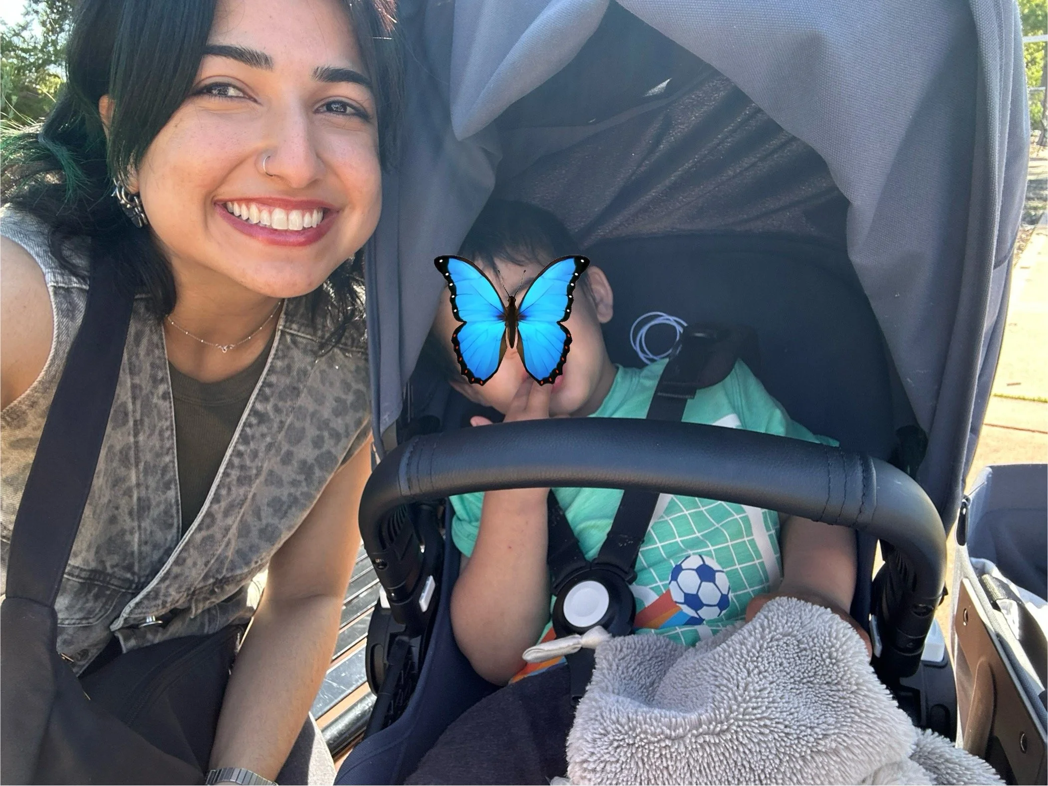 A smiling woman with dark hair, earrings, and a nose ring leaning into a stroller. The stroller contains a young child whose face is covered by a large blue butterfly graphic overlay.