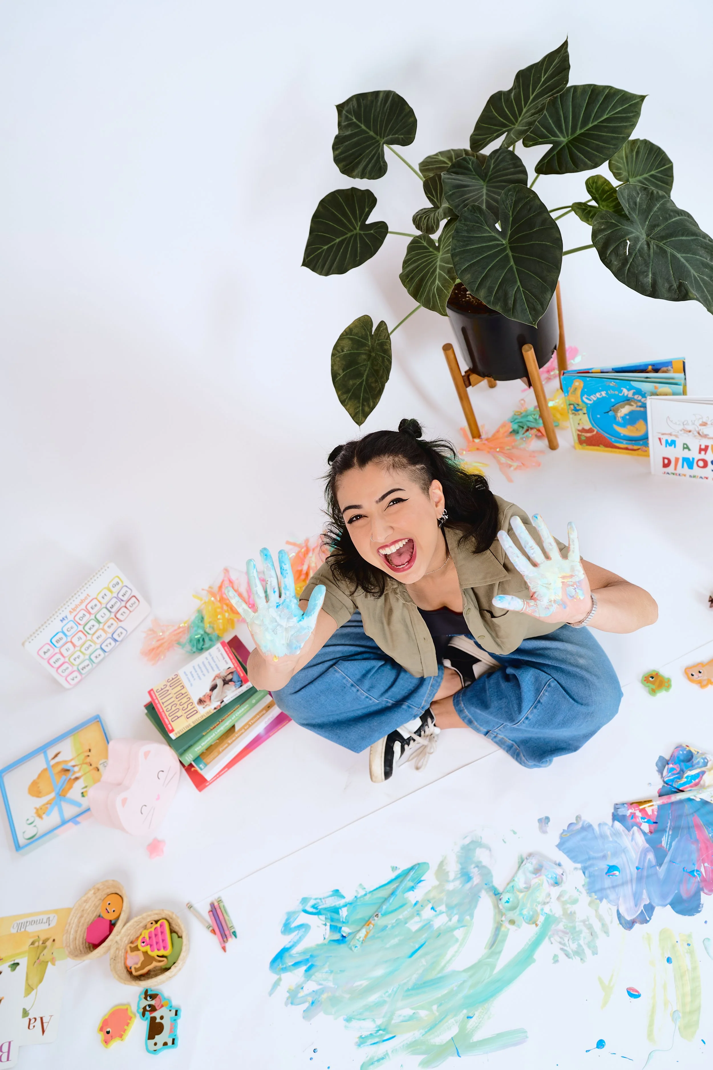 A young woman with dark, curly hair in pigtails, smiling and showing her hands covered in colorful paint, sitting on a white floor with children's books, toys, and art supplies around her, with a large green plant in a pot nearby.