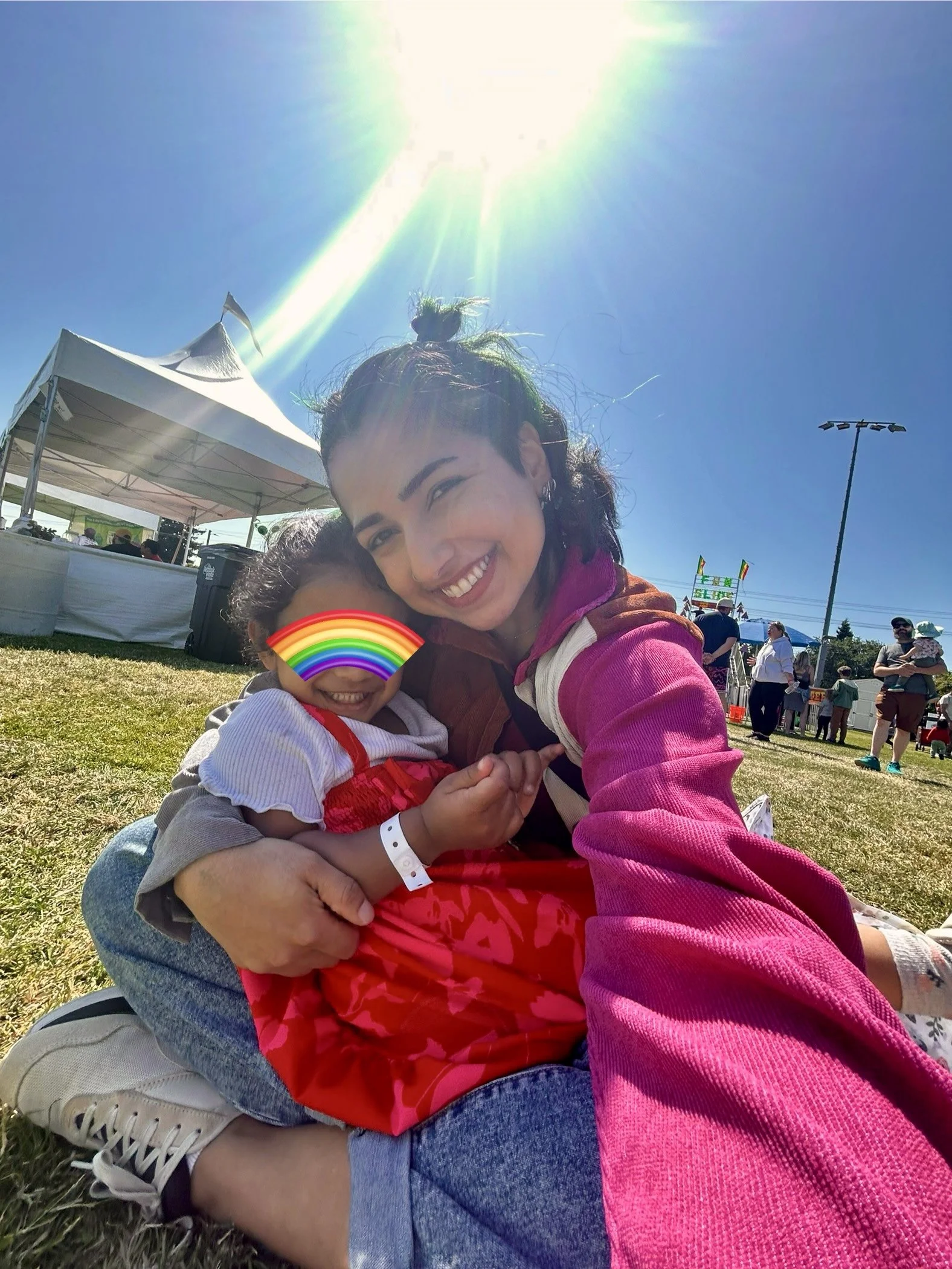 A woman with curly dark hair tied in a bun, smiling and hugging a young girl. The girl has a rainbow sticker covering her face, is wearing glasses, and a white hoodie. They are outside under a bright sun, with white tents and people in the background at a sunny outdoor event.
