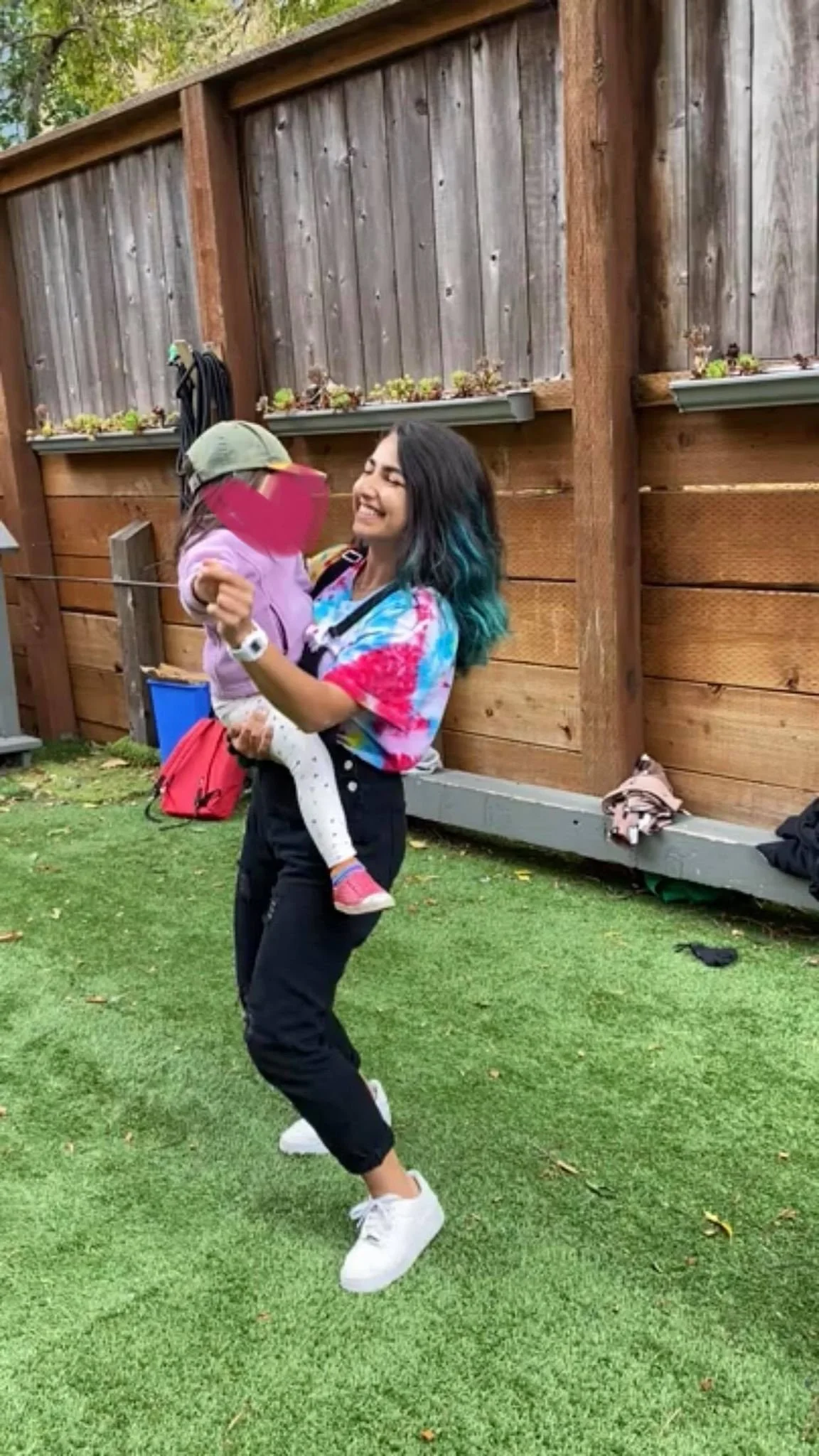 A woman smiling and dancing outdoors while holding a young girl in her arms, with a wooden fence and green grass in the background.