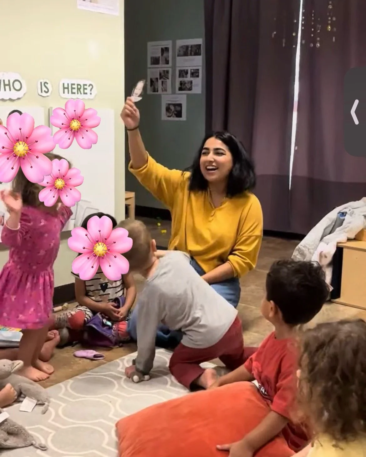 A woman in a yellow shirt sitting on the floor surrounded by children with pink flowers covering their faces in a classroom setting, engaging in a group activity or storytelling.