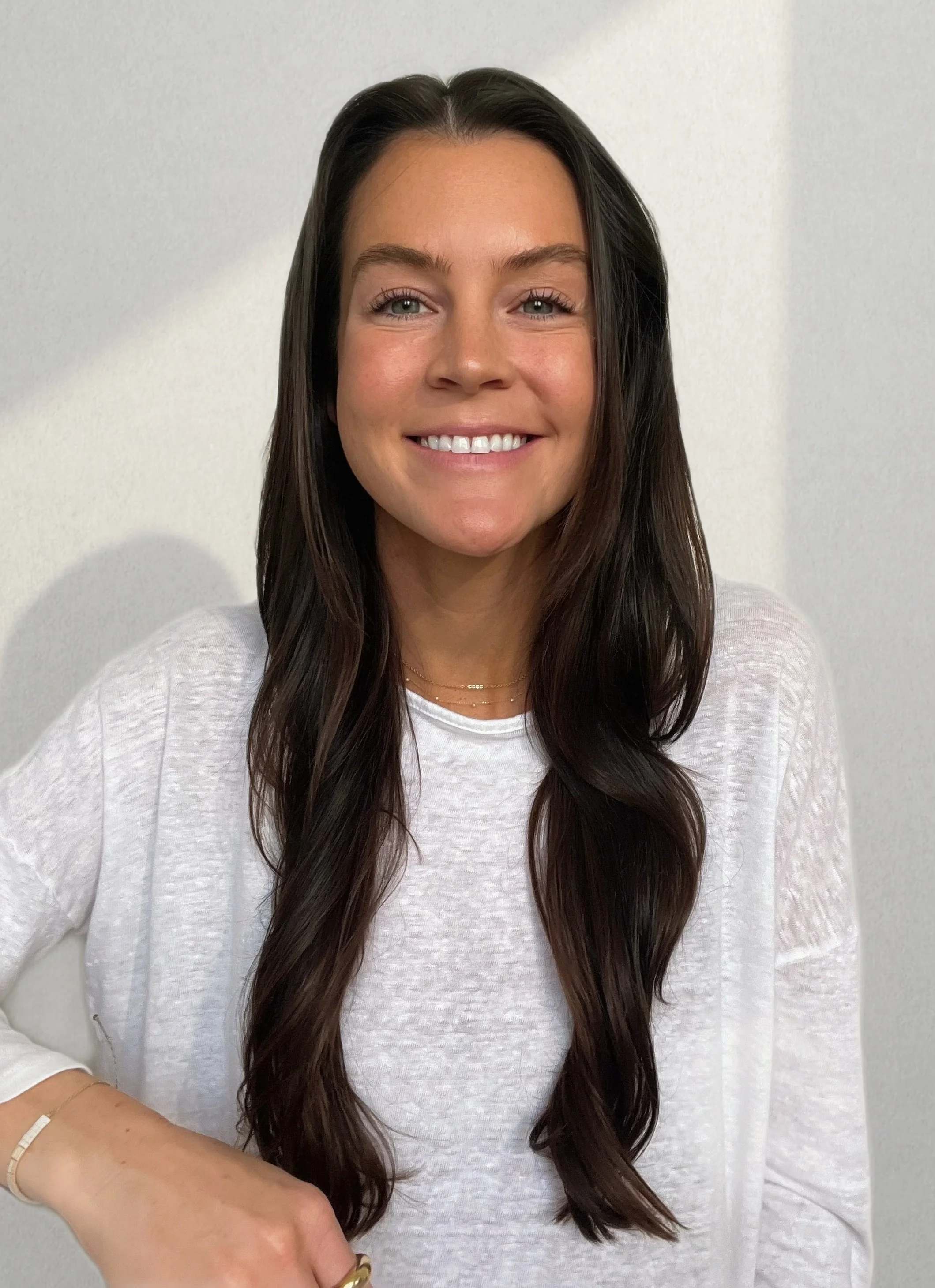 A woman with long, dark hair, wearing a white top and jewelry, smiling in front of a plain light-colored wall.
