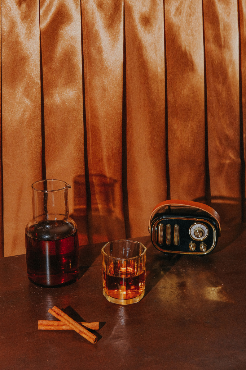 A still life scene featuring a glass flask filled with red liquid, a short glass tumbler with a similar beverage, two cinnamon sticks, a vintage styled radio, and a brown curtain in the background.