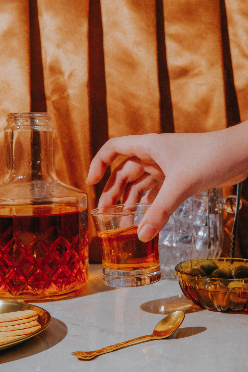 A hand is reaching to pick up a glass of amber-colored liquid from a table, with a decanter of the same liquid, an empty glass, a bowl of pickles, a plate of crackers, and a gold spoon also on the table.