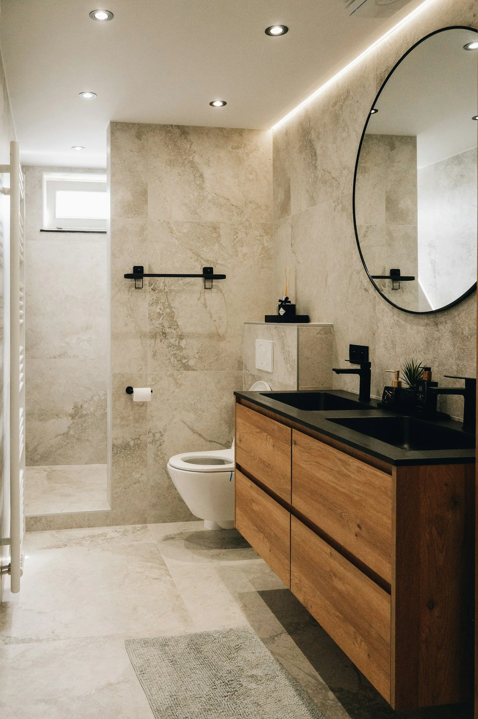 Modern bathroom with beige marble walls and floor, a wooden vanity with black countertop and dual sinks, a round mirror, a black faucet, a white toilet, a small window, wall shelves, and recessed ceiling lights.