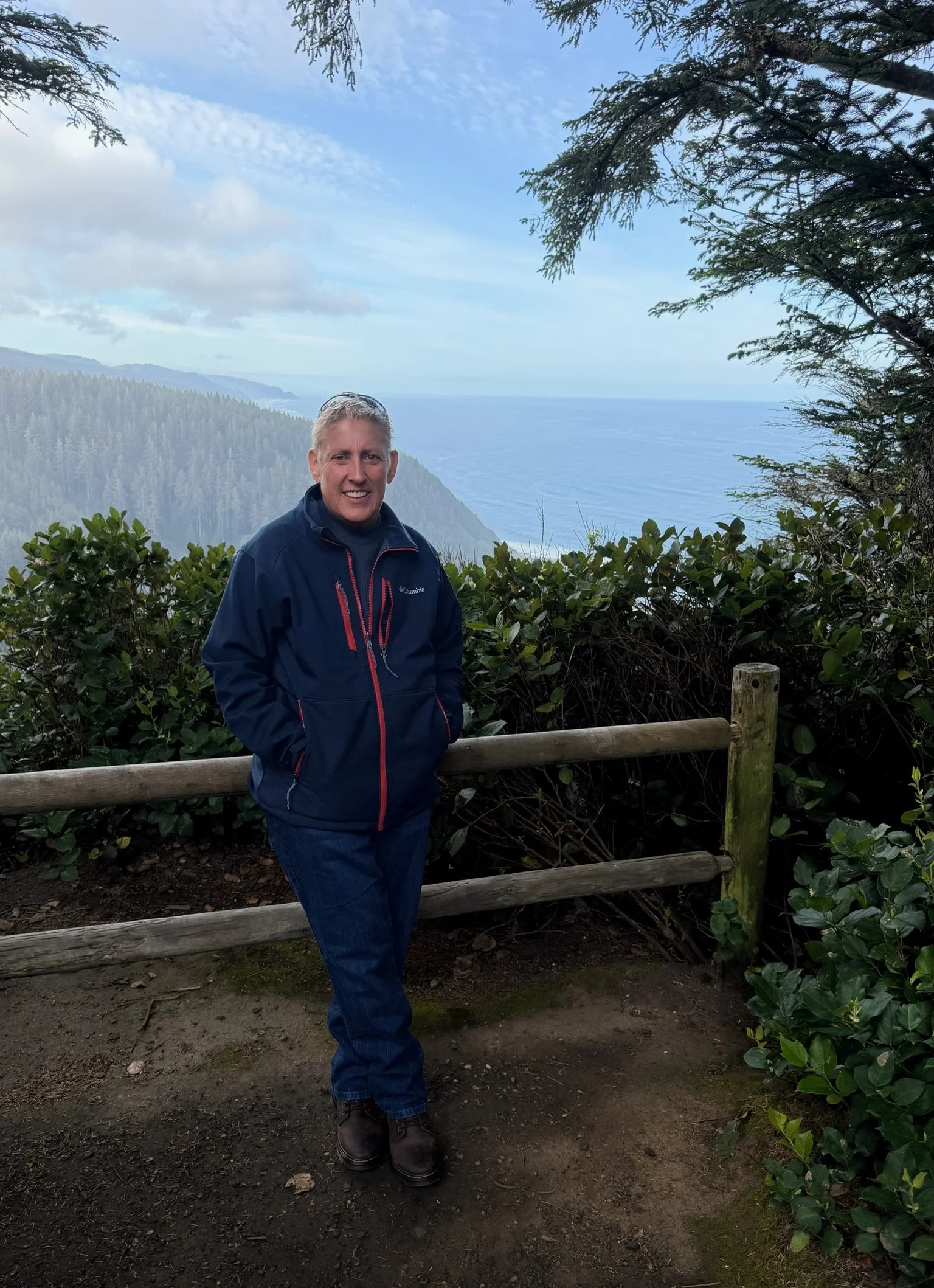 A man standing outdoors on a dirt path behind a wooden railing, with lush bushes, trees, and a scenic view of hills and ocean in the background.