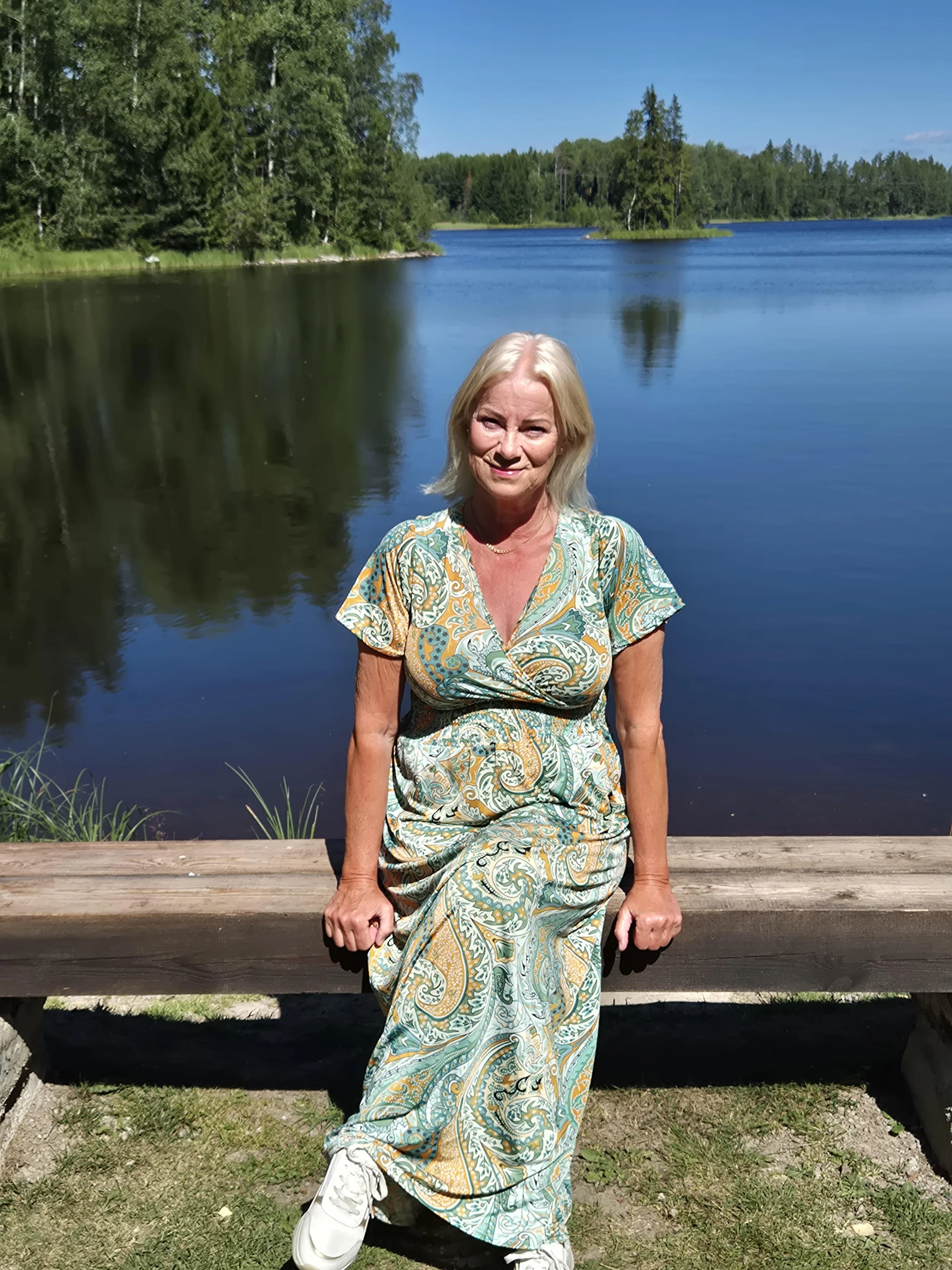 En kvinna i en sommarklänning som sitter på en träbänk vid en sjö, omgiven av gröna träd och ett klart blått himmel.