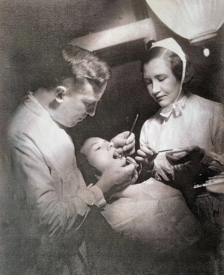 Svartvit bild av en barnläkare som undersöker en pojke medan en sjuksköterska hjälper till på ett klinikrum.