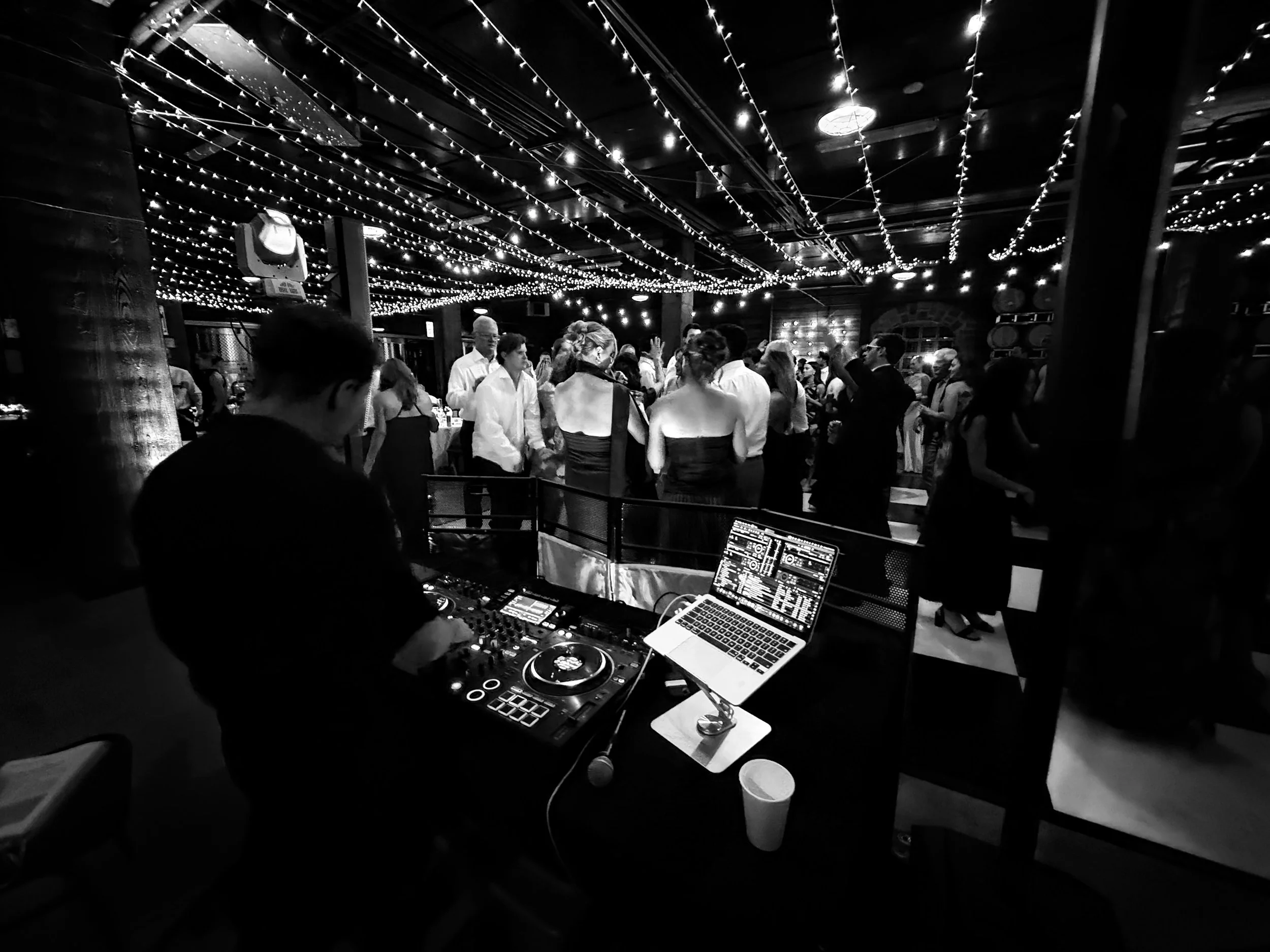 A DJ is performing at a crowded indoor party with string lights hanging from the ceiling.