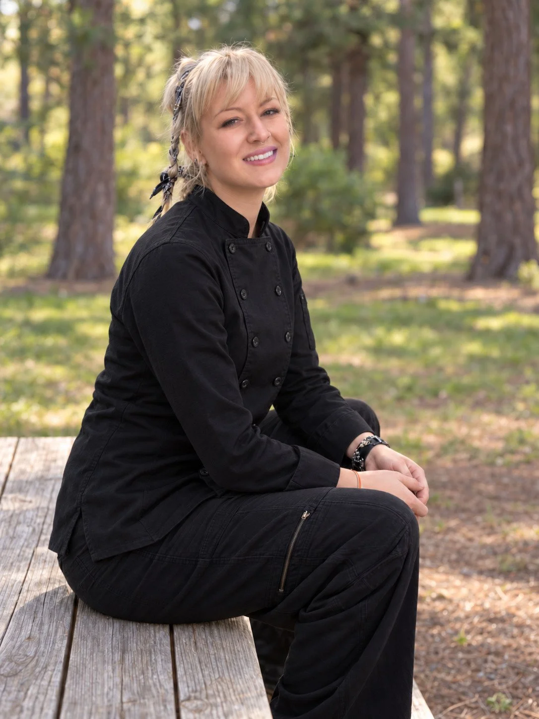 A woman in a black chef's jacket sitting on a wooden bench outdoors in a park with trees in the background.
