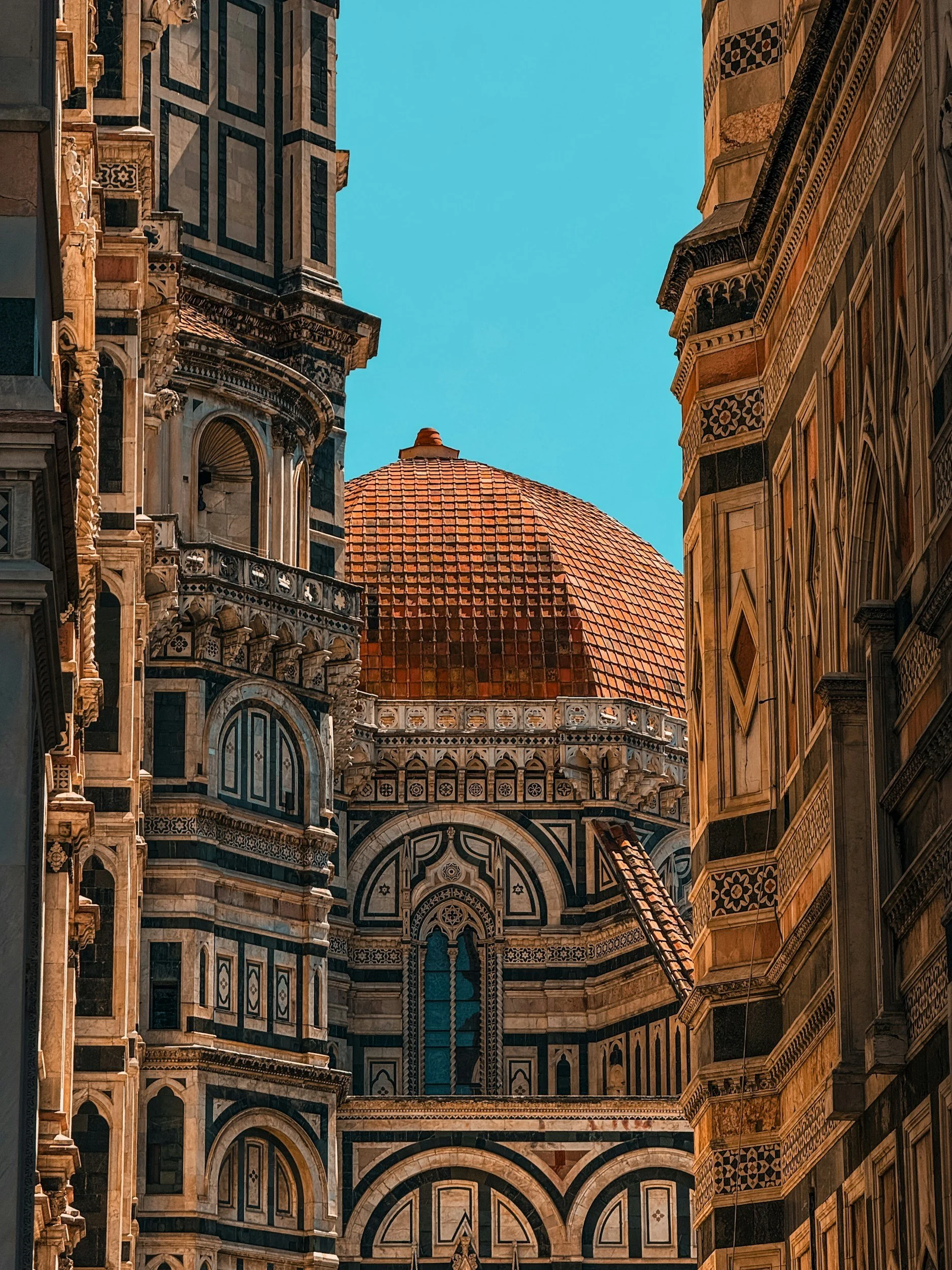 View of the ornate marble architecture of Florence, Italy Cathedral with a large red-tiled dome in the background under a clear blue sky. The Duomo.