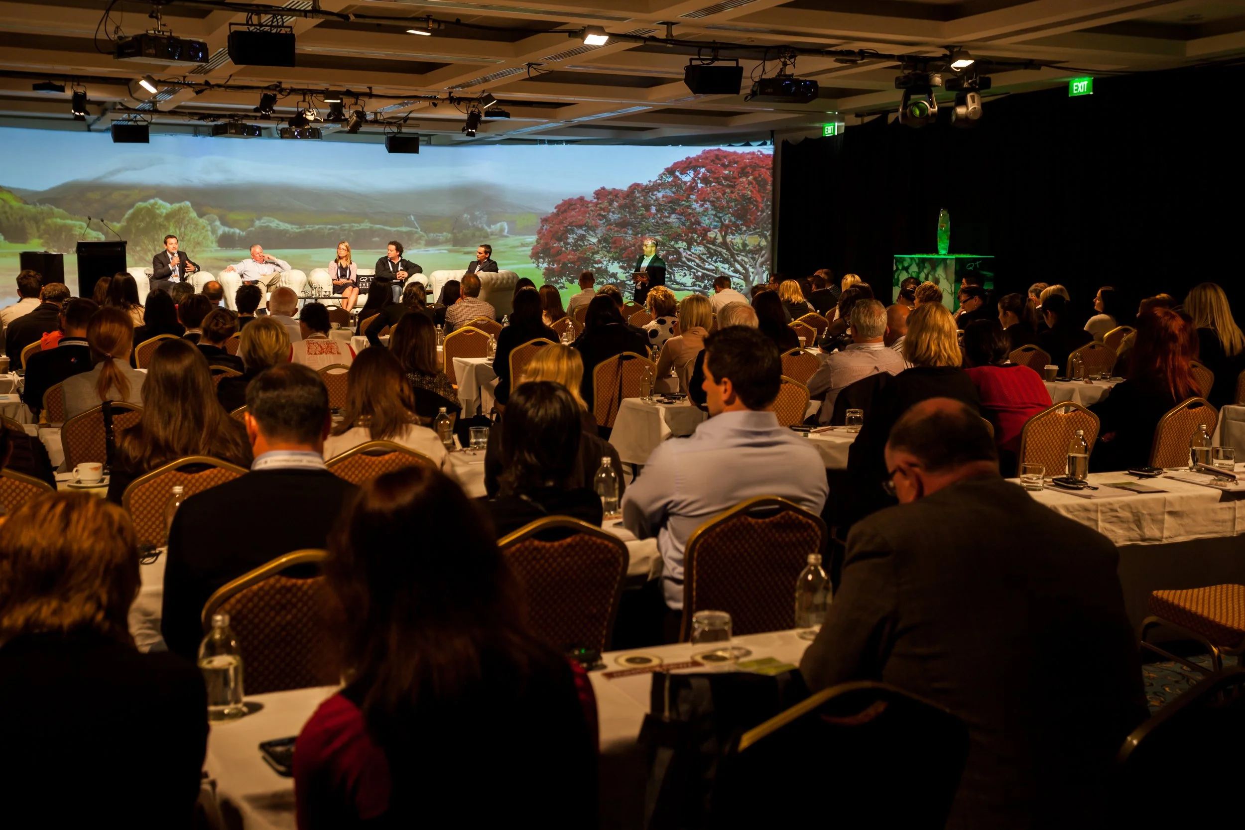 A large conference room filled with round tables and many attendees watching a panel discussion on stage. The stage features four panelists seated on curved white couches and one person standing at a podium. A large scenic backdrop with trees and a landscape is visible behind the stage. The room is well-lit with ceiling lights and spotlights.