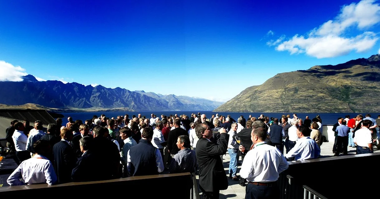 A large group of people gathered outdoors by a lake with mountains in the background on a clear day