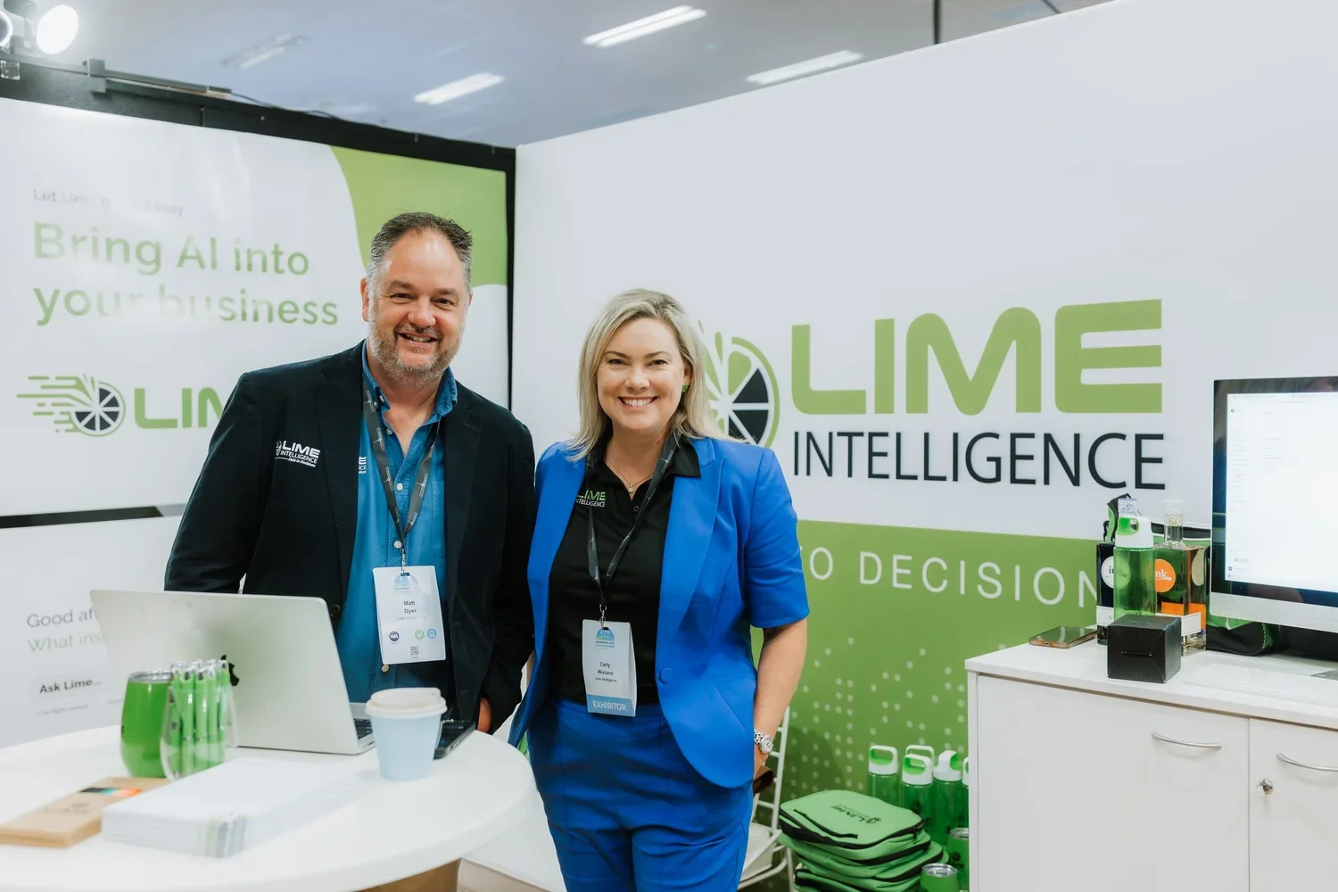 Two people, a man and a woman, standing at a trade show booth with Lime Intelligence branding. The man is wearing a black blazer and blue shirt, and the woman is wearing a blue blazer and black shirt. Both are smiling and wearing exhibitor badges. The booth has a green and white color scheme, with promotional materials, a laptop, water bottles, and a computer monitor visible.