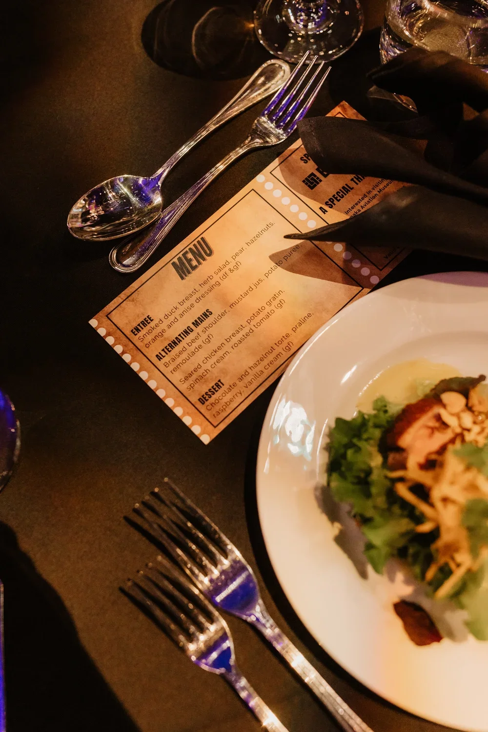 A table setting with a knife, fork, and spoon, a menu card, a partial view of a salad on a white plate, and glasses of water on a dark table surface.
