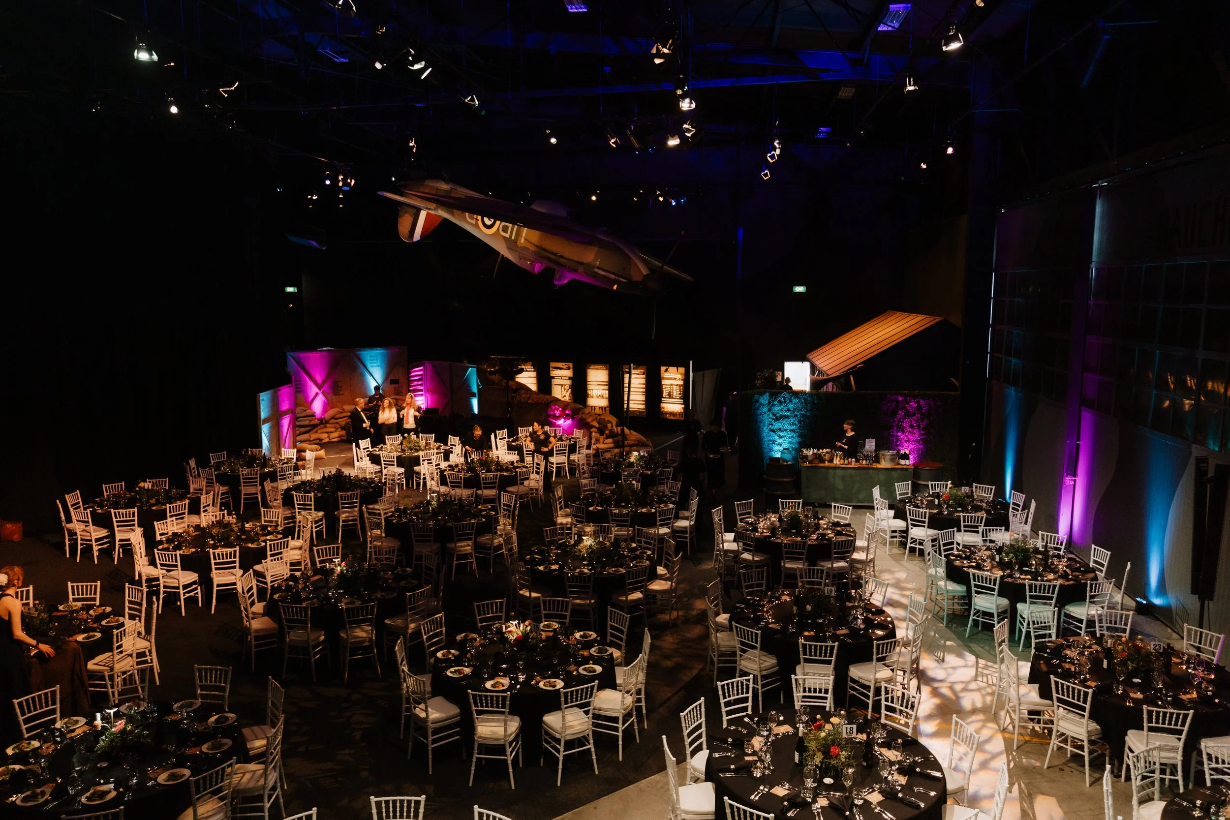 An event space set up with round tables and white chairs, decorated with flowers, with a jazz aircraft on a ledge and colorful wall lighting, and some people preparing for the event.