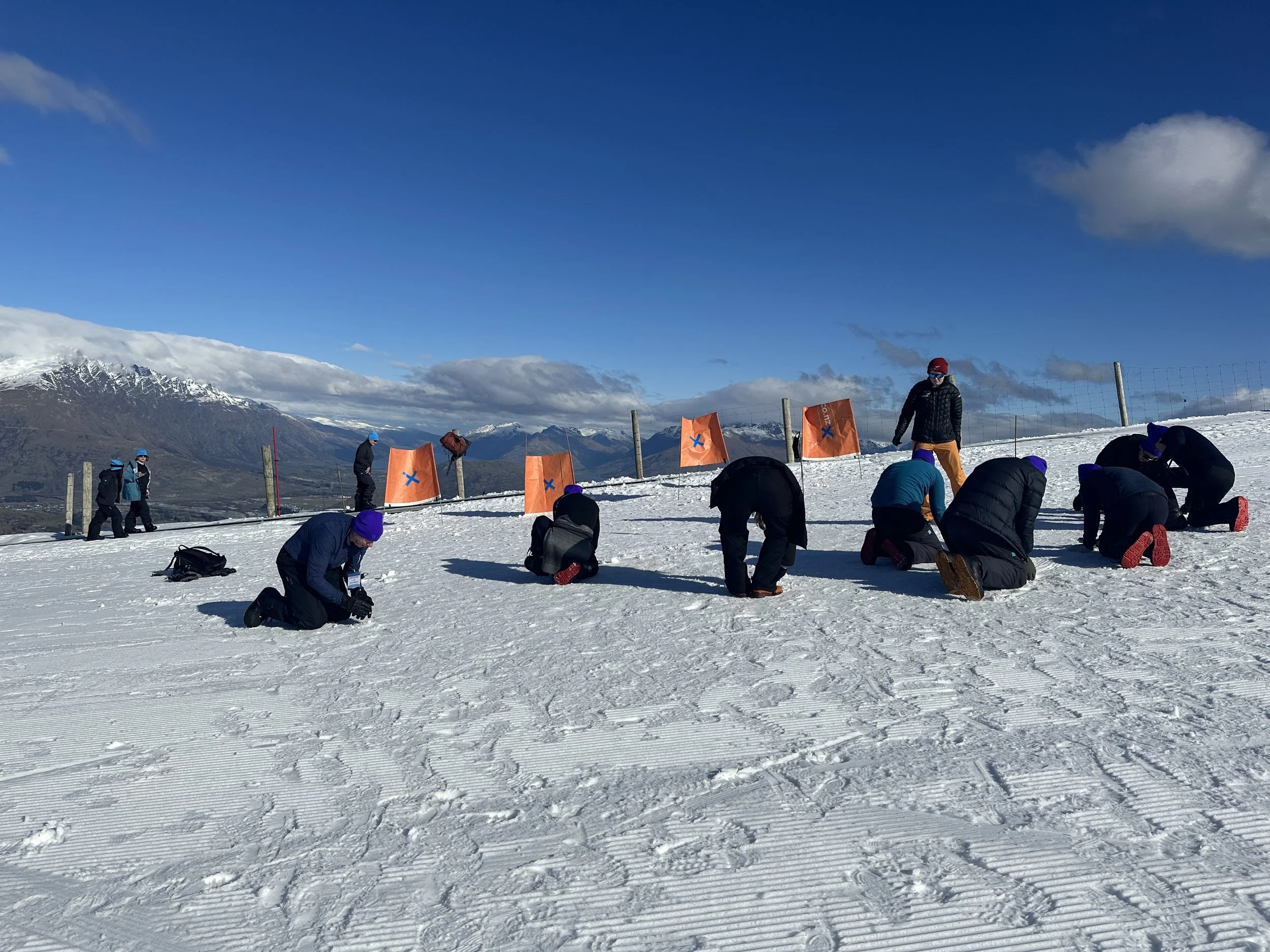 People on a snowy mountain slope, some kneeling and others standing, with bright orange flags and snowy mountains in the background under a blue sky with a few clouds.