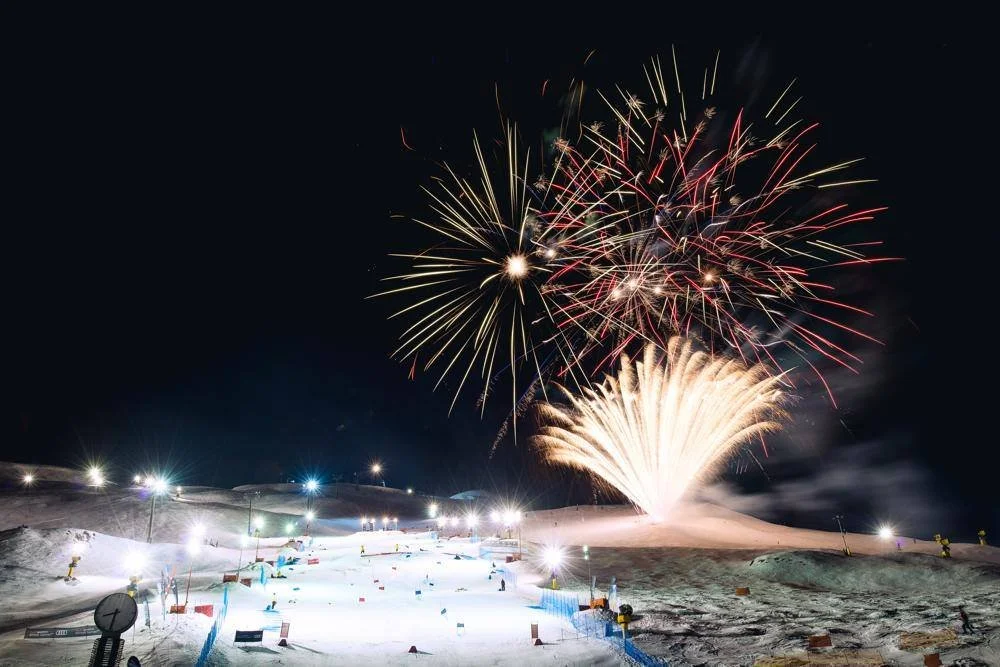 Fireworks exploding in the night sky over a snow-covered landscape with ski slopes and bright lights.