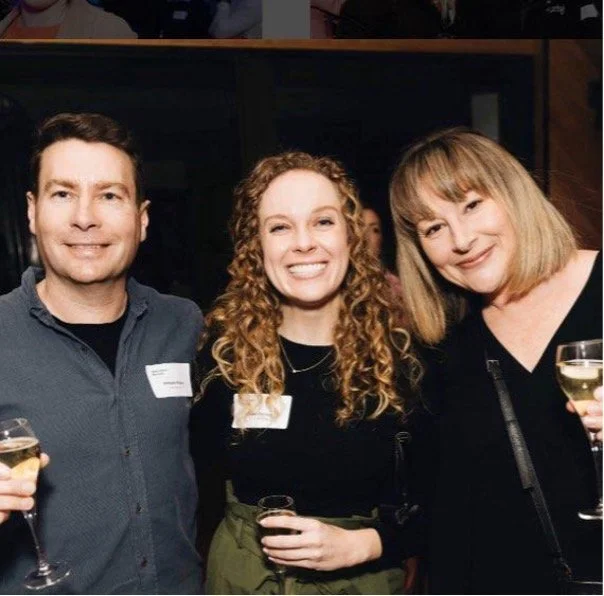 Three people smiling at a social event, holding glasses of white wine.