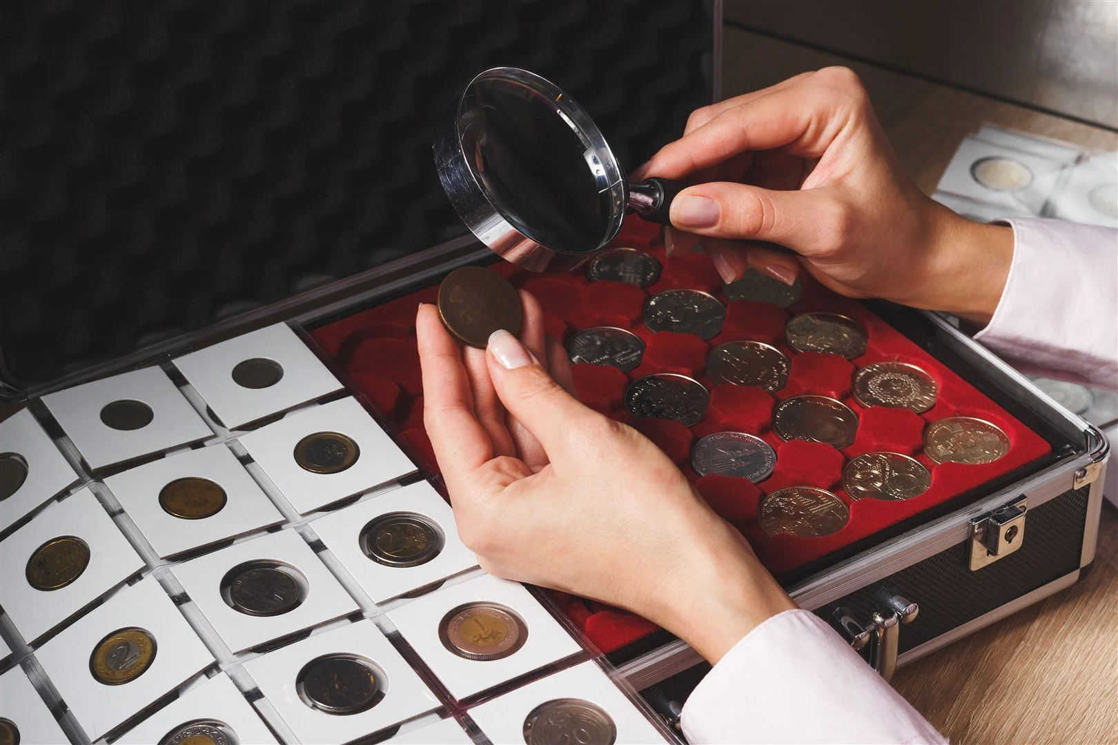 Person holding a magnifying glass inspecting euro coins in a red-lined case.