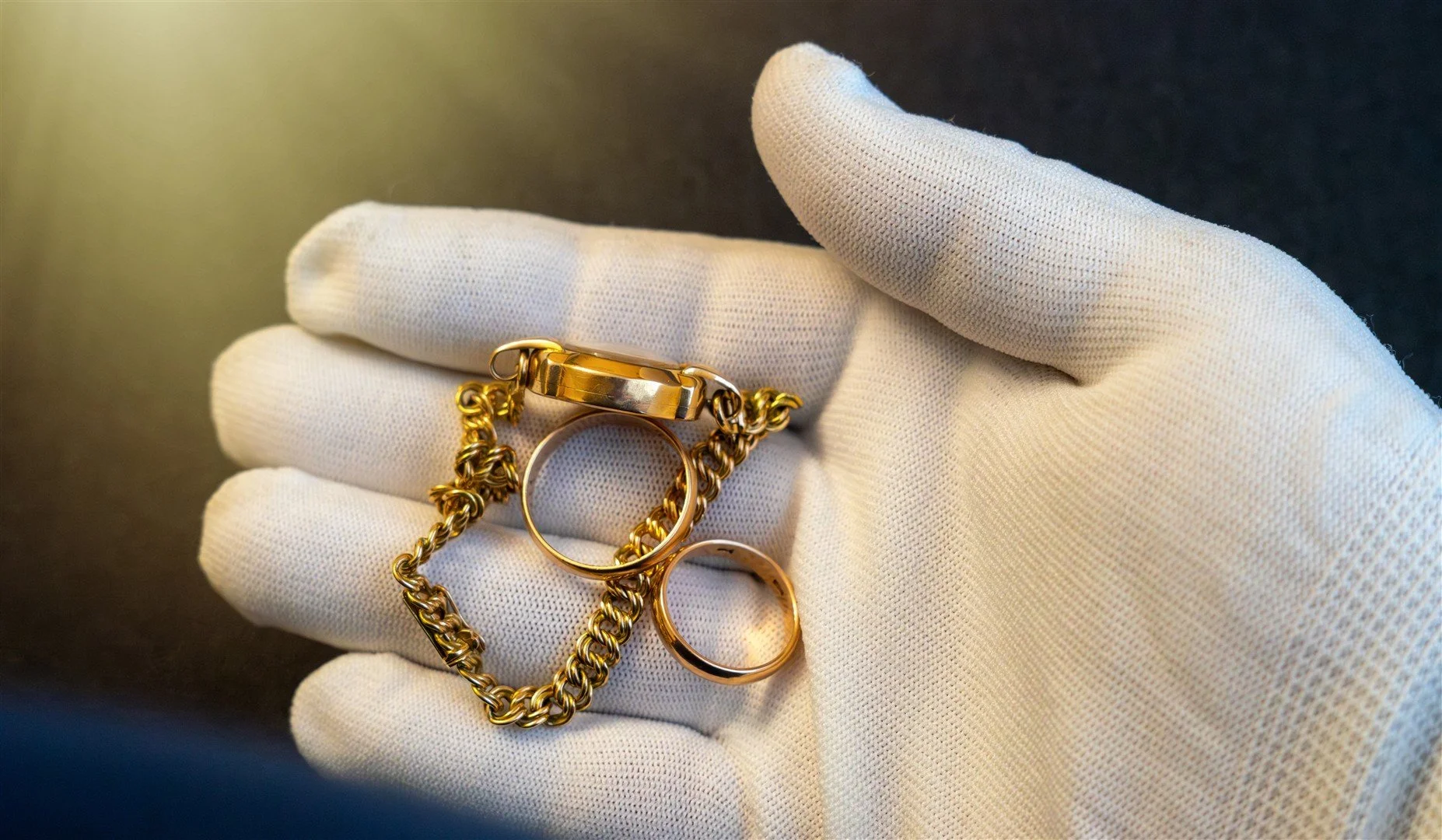 A gloved hand holding a collection of gold jewelry including rings, a chain, and a bracelet.
