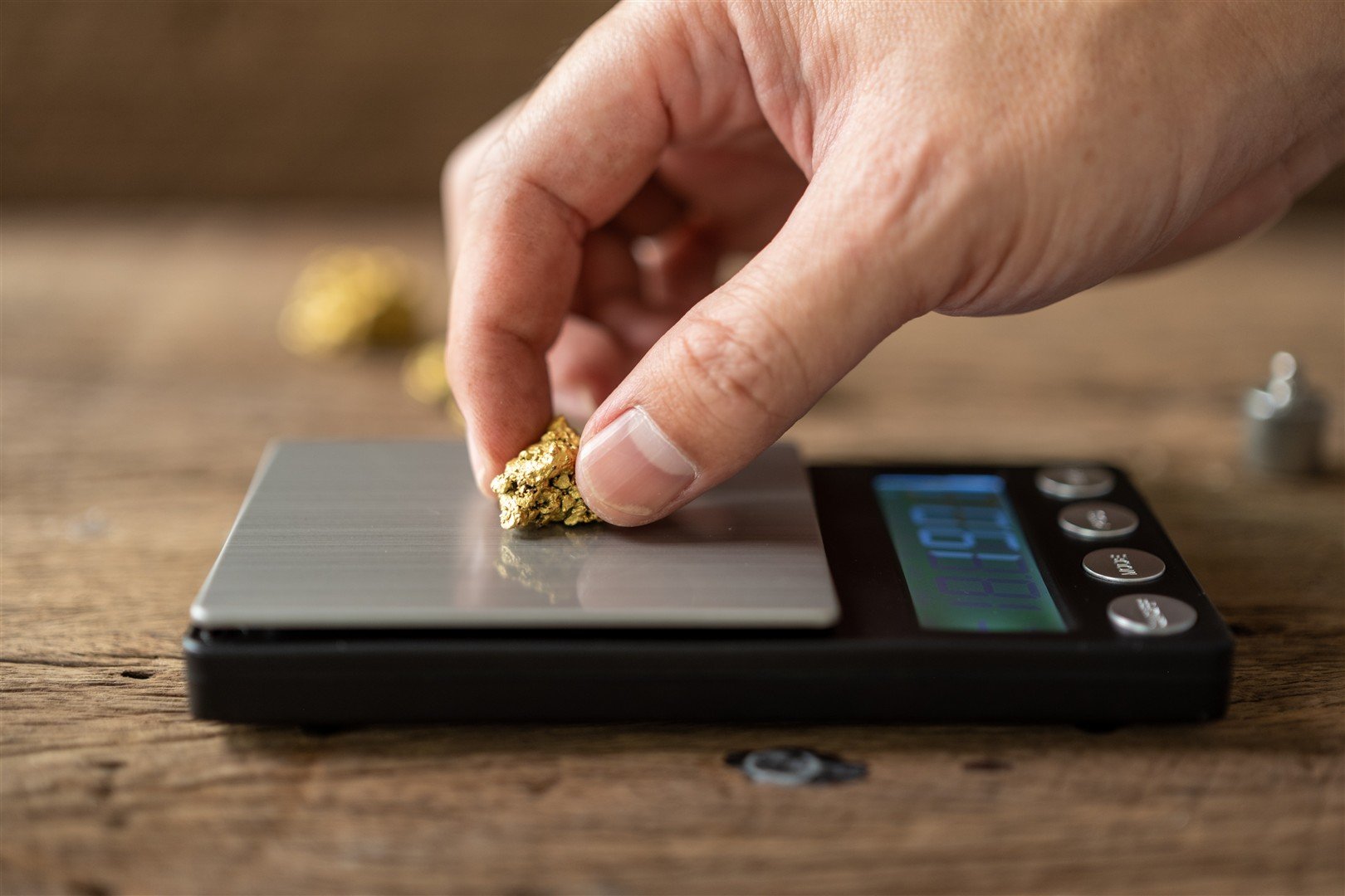Hand weighing a small gold nugget on a digital scale on a wooden surface.