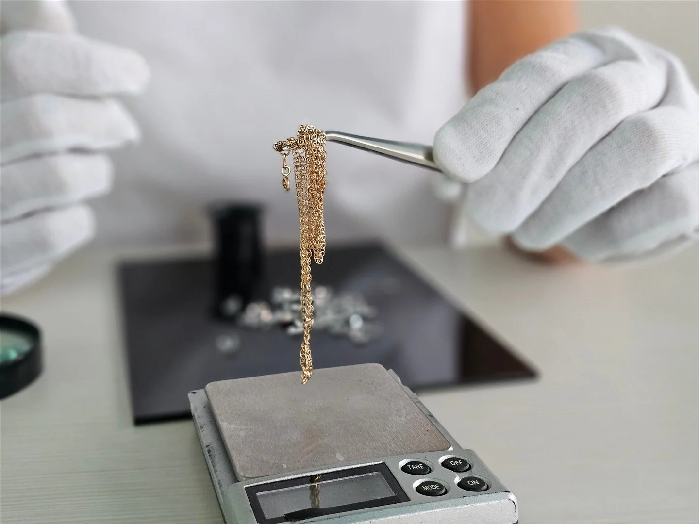 Person wearing white gloves holding a gold chain with jewelry tools, with a digital scale in the foreground, jewelry workspace in the background.