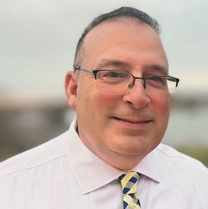 Josh Deering, a white man wearing rectangular half-frame glasses, a professional white button down shirt, and a yellow and blue neck tie.