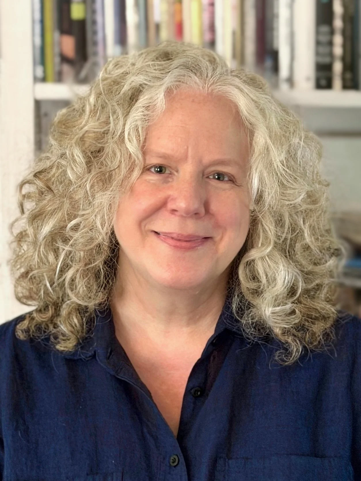 Kim Simon, Registered Psychotherapist (Qualifying), with curly blond-grey hair and a soft smile, wearing a blue shirt, posed in front of a bookshelf.