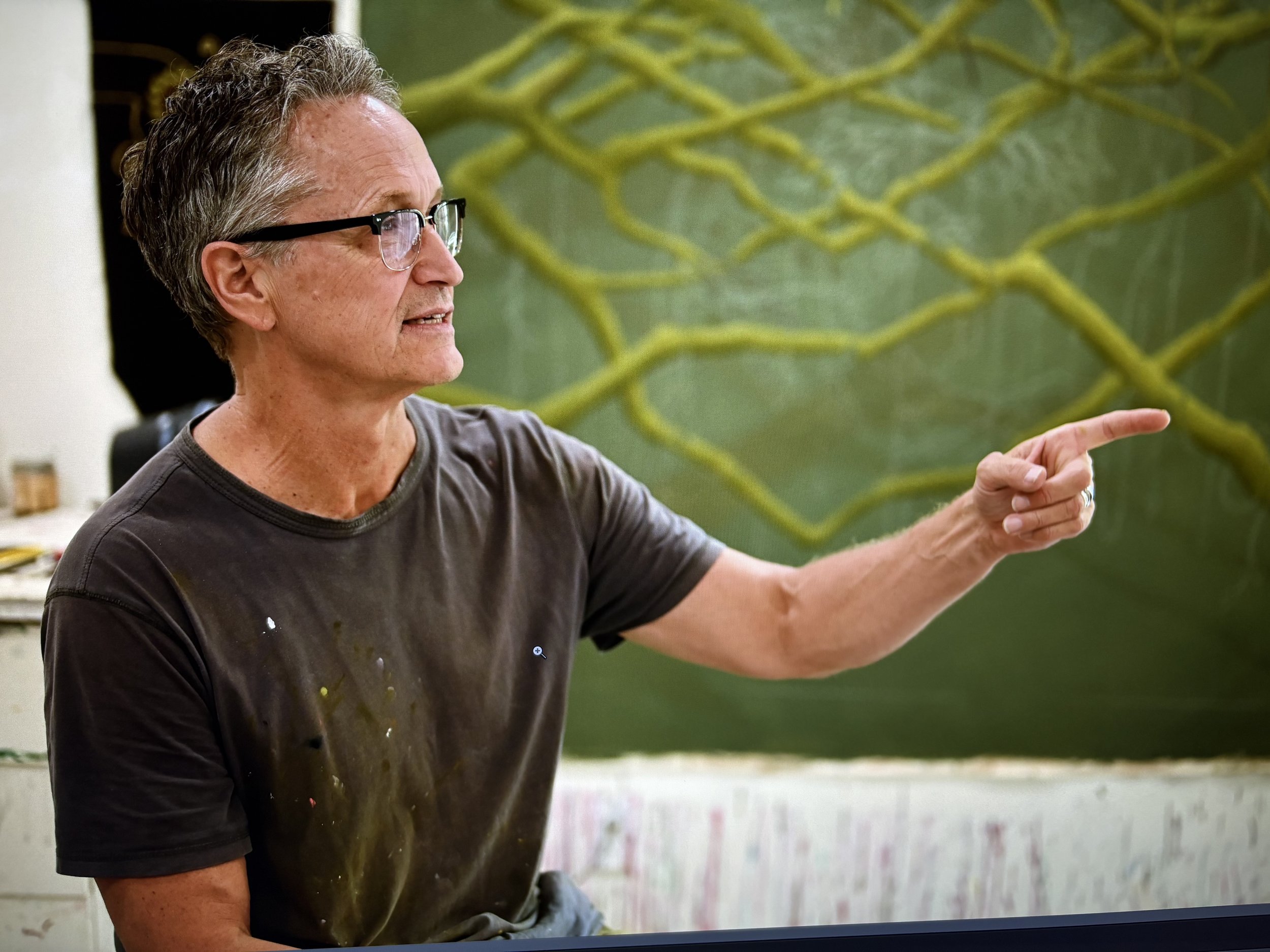 A man with glasses and gray hair pointing at something. He is wearing a gray T-shirt with paint stains, standing in front of a green wall with grass or plant design.