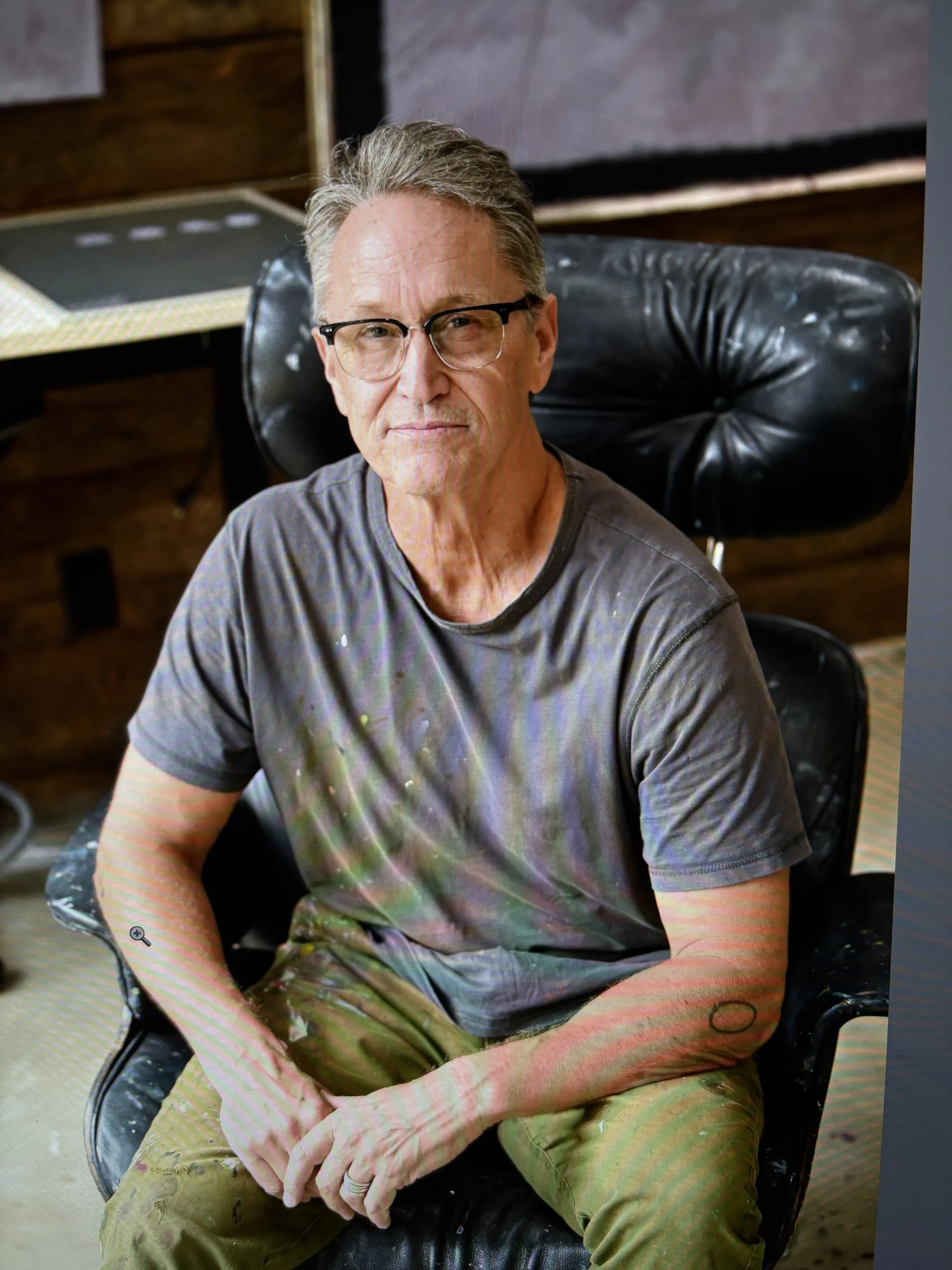 A man with gray hair and glasses sitting in a leather chair. He is wearing a gray t-shirt and green camouflage pants, with some paint stains. The background includes a wooden wall, a table with a laptop, and colored vertical curtains.