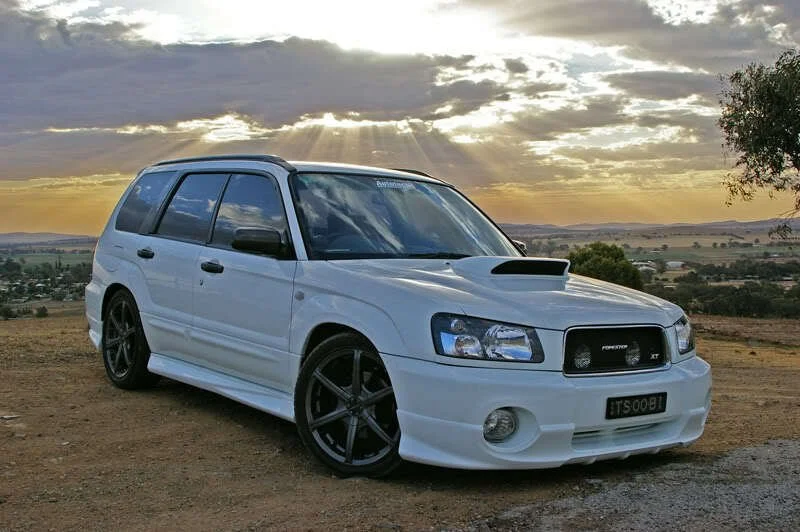 A white Subaru Forester XT with black custom wheels, parked on a dirt road at sunset with a rural landscape in the background.