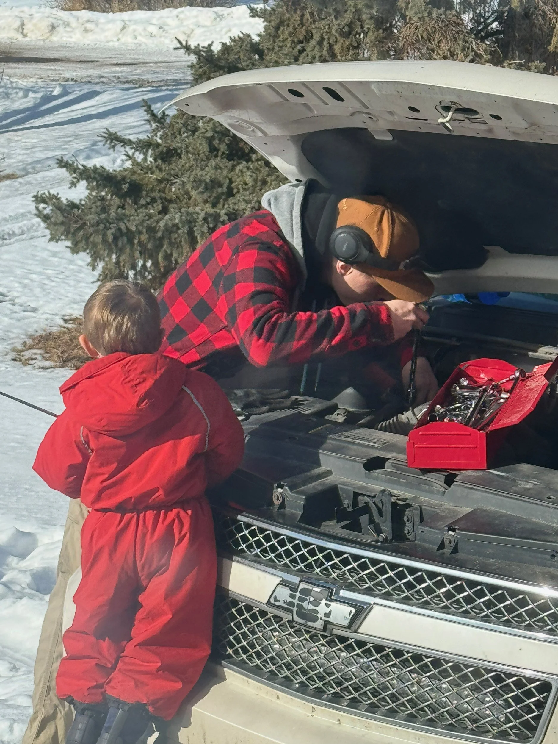 Two children in winter clothing working on a car engine with the hood open in a snowy outdoor setting, with trees and snow-covered ground in the background.