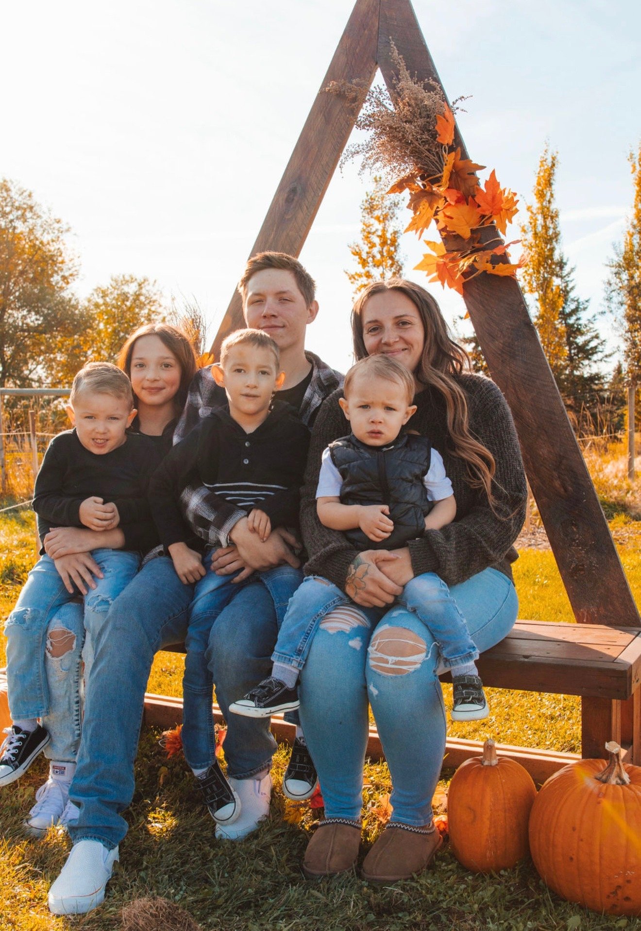 A family of six posing outdoors during fall with pumpkins and autumn foliage in the background.