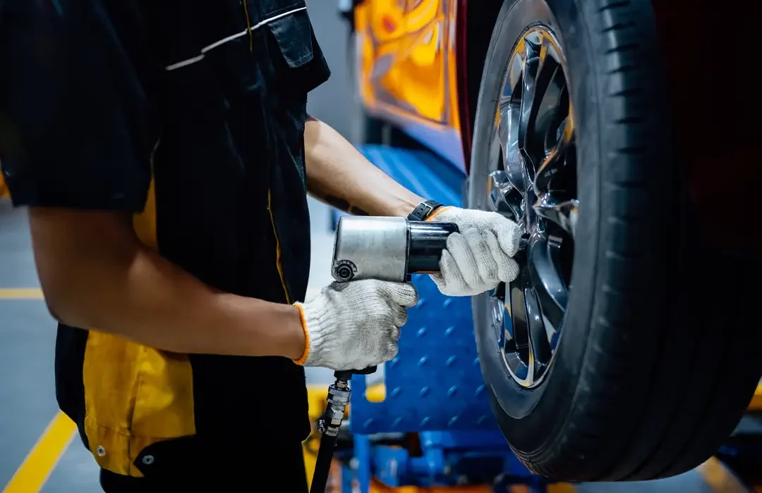 A mechanic wearing gloves is using a pneumatic impact wrench to tighten or loosen bolts on a car tire in an auto repair shop.