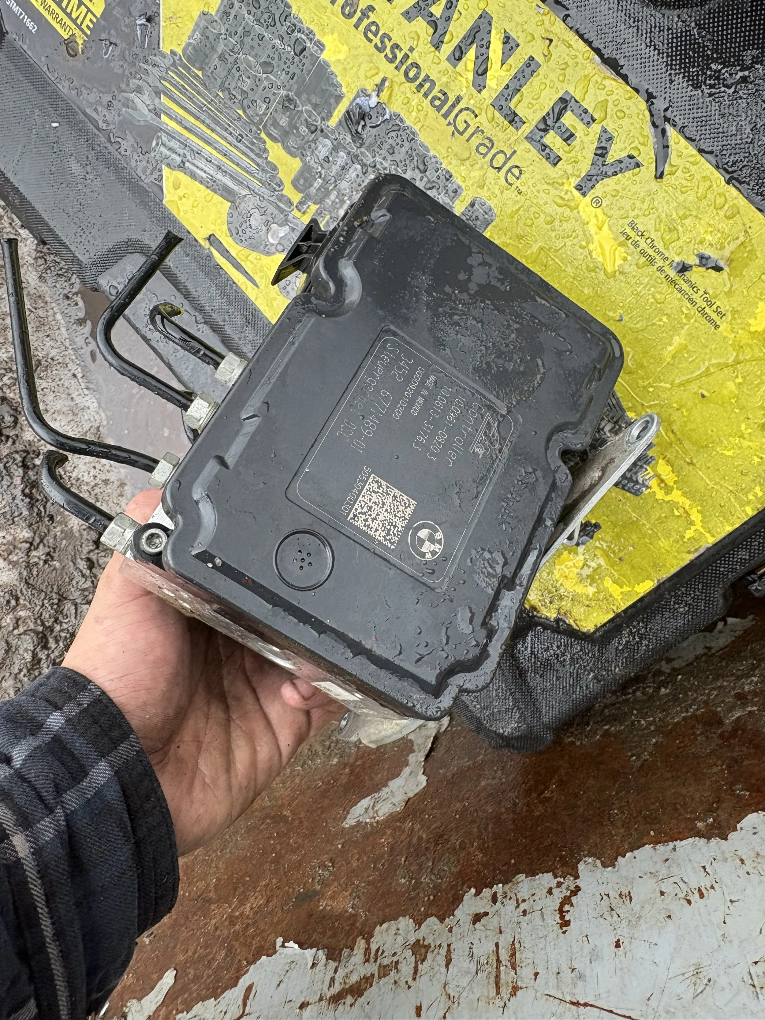 Close-up of a mechanical device, likely an electronic control module, held in a person's hand against a background of a yellow work surface with parts and tools for automotive repair.