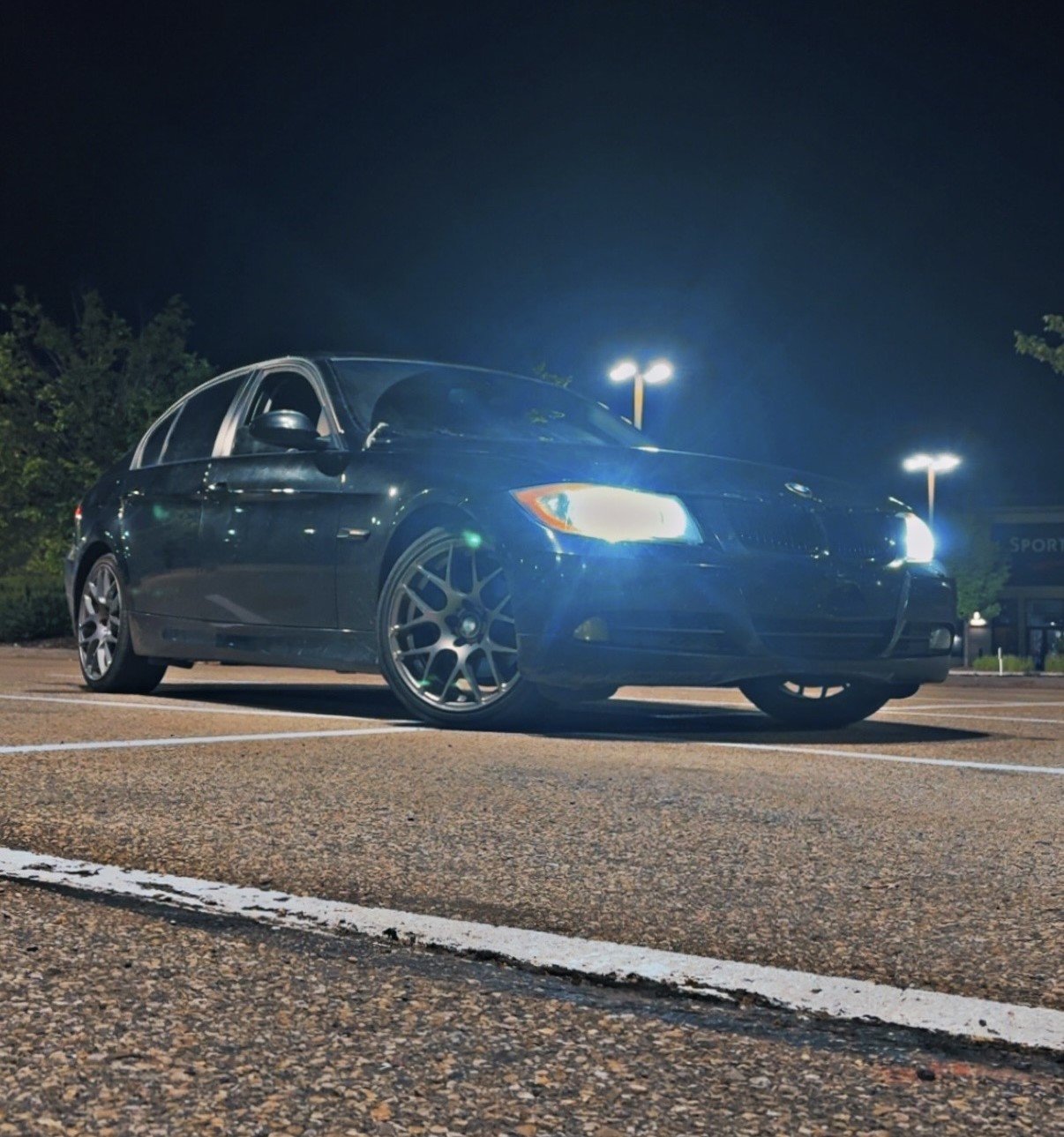 A black sedan car parked in a parking lot at night with its headlights on and streetlights illuminating the area.