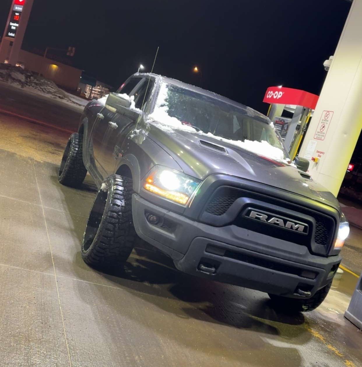 A black RAM pickup truck parked at a gas station at night with snow on the hood and windshield.