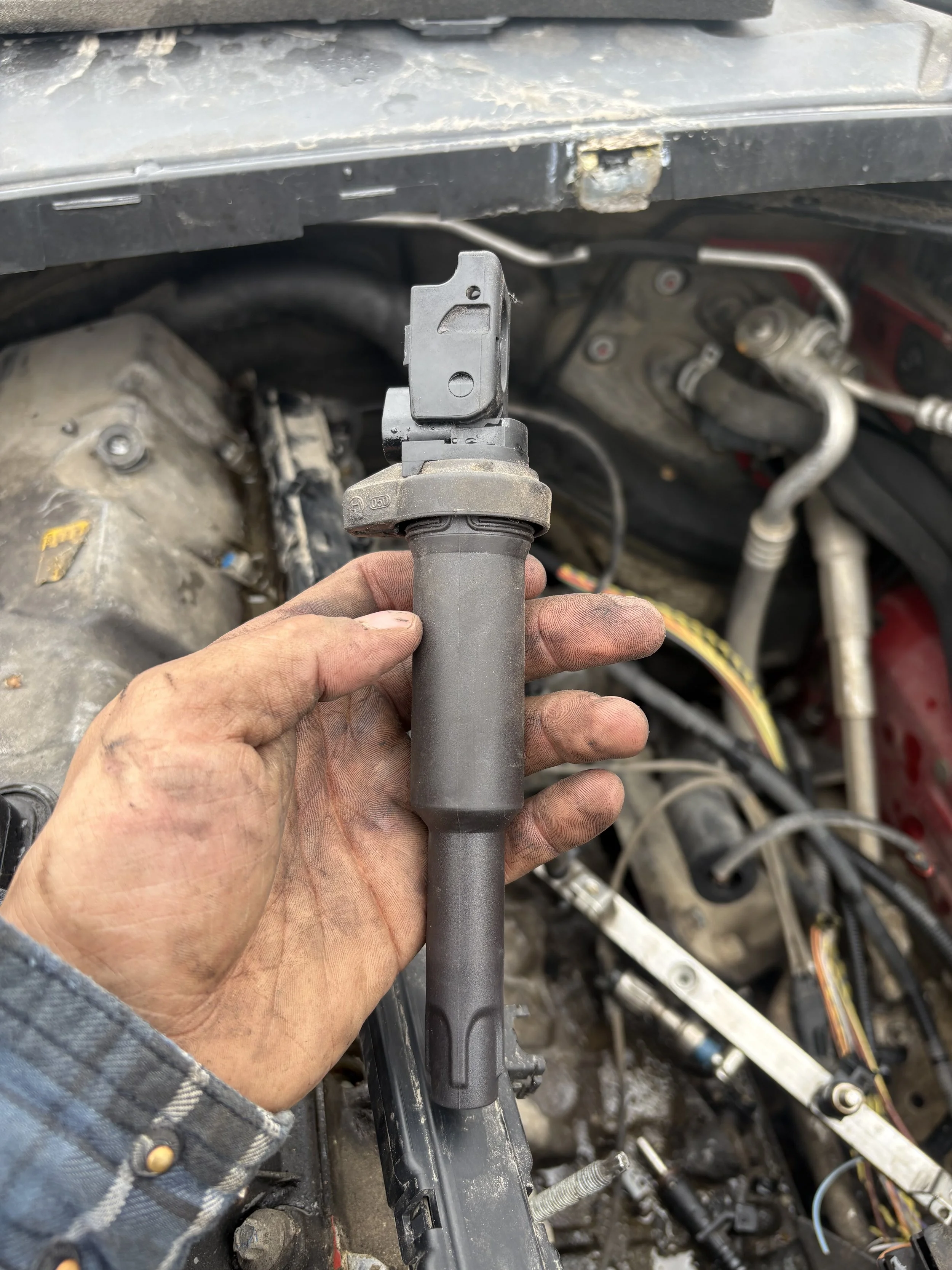 A person holding a specialized metal tool with a hook and slot, working under the hood of a car with visible engine components, hoses, and wires.