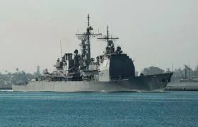 Military battleship sailing in the water with a cityscape in the background.