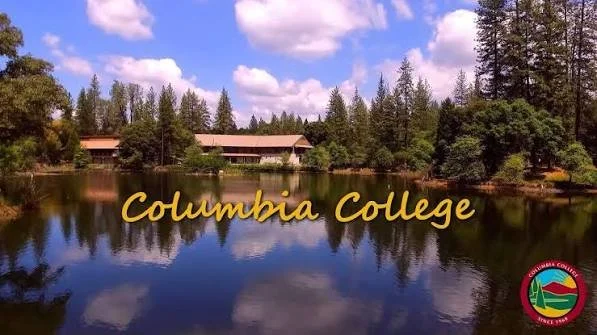 A serene lake surrounded by trees with a clear blue sky and scattered clouds, featuring the text 'Columbia College' in yellow